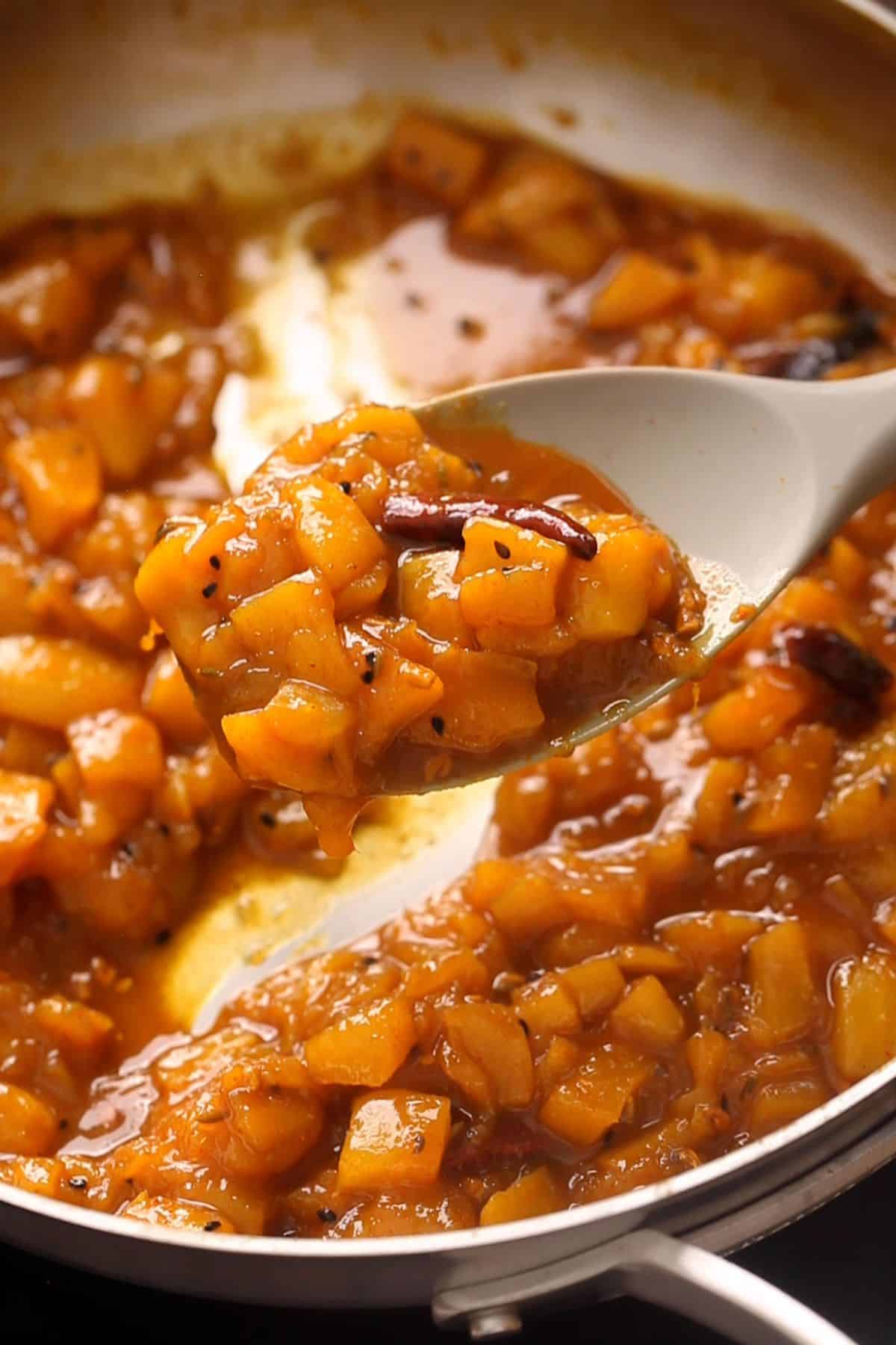 A close-up of a pan filled with chunky mango chutney. The chutney is a rich brown color, with visible pieces of mangoes and spices.