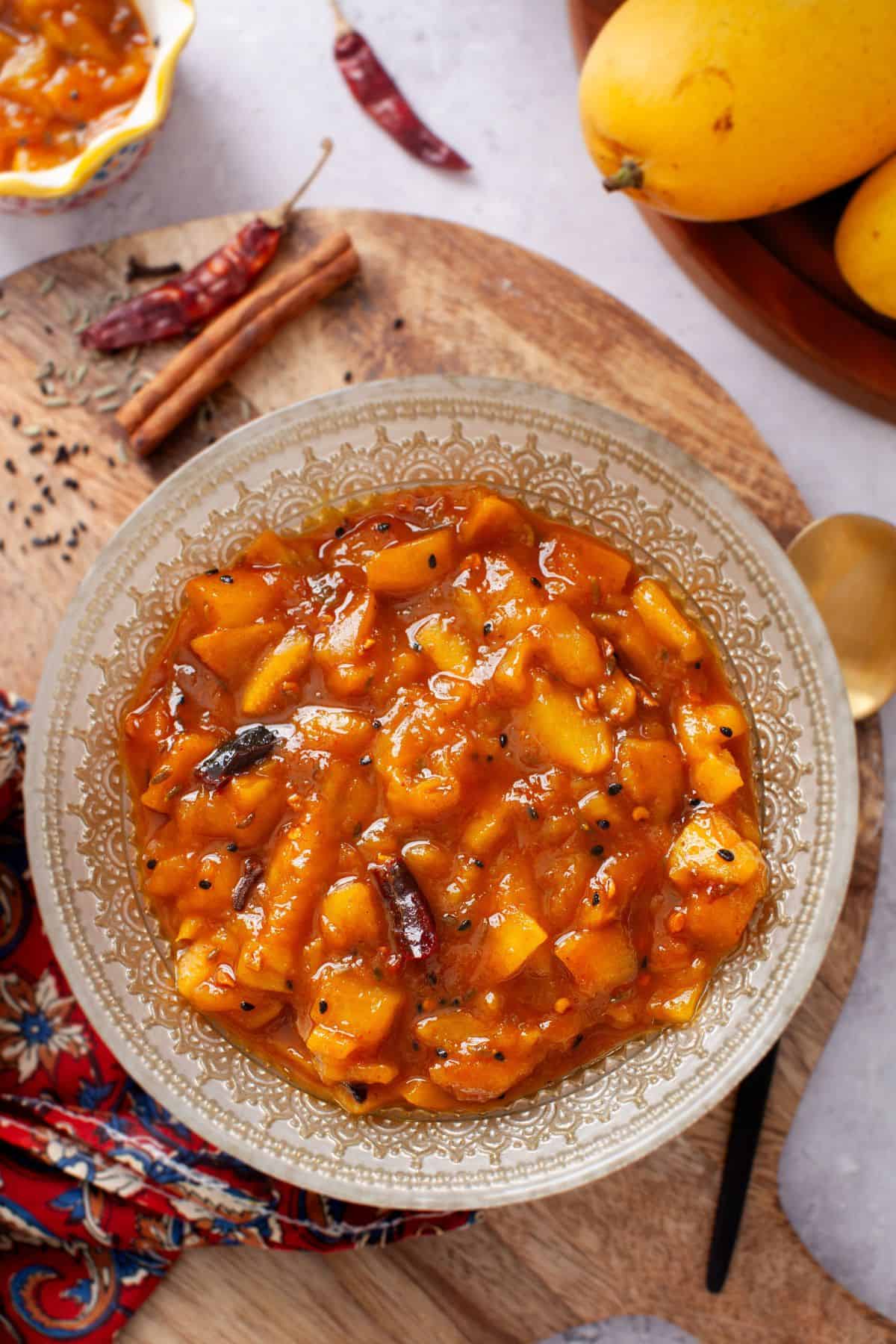 A bowl of mango chutney on a decorative plate, surrounded by spices like red chili, cinnamon sticks, and whole mustard seeds.