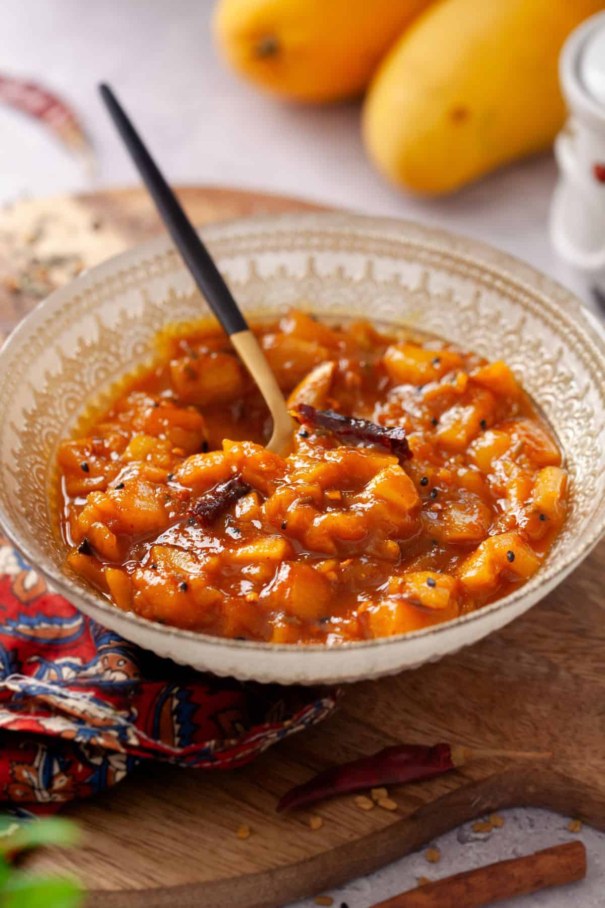 A bowl of mango chutney with a spoon, placed on a wooden board. The chutney is vibrant orange with visible chunks of mango and seeds.