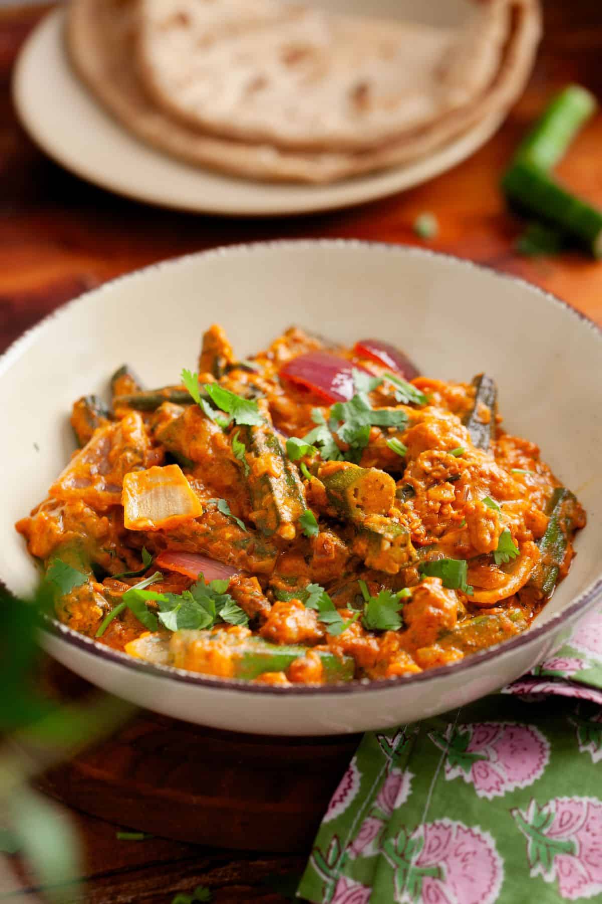 A bowl of Indian bhindi masala garnished with fresh cilantro, with a plate of chapati in the background.