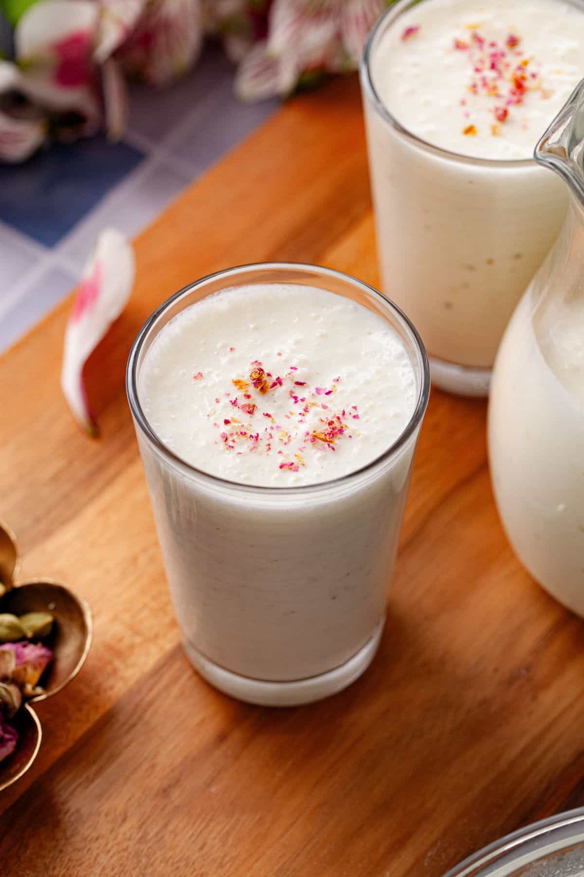 Creamy white sweet lassi topped with rose petals in a glass, placed on a blue & white tiled table.