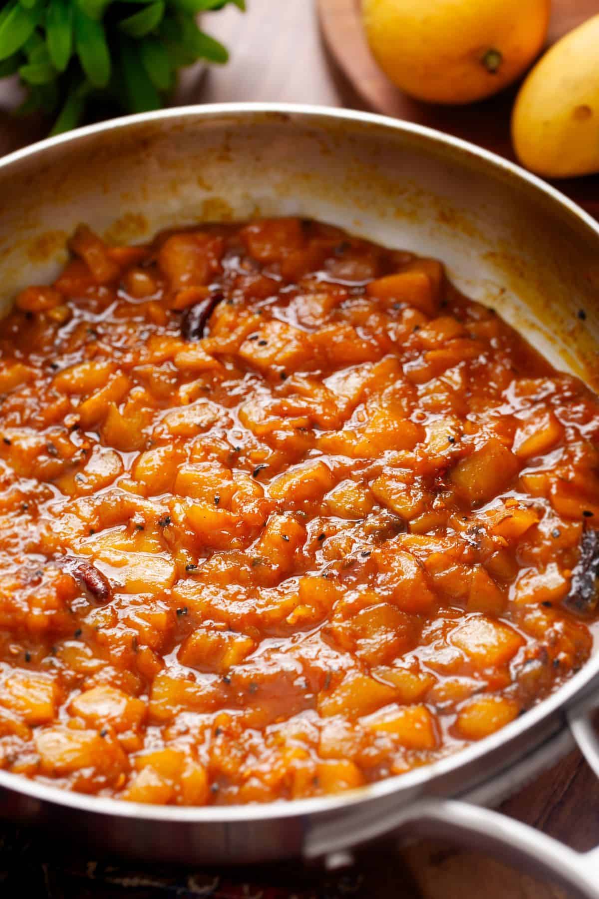 A close-up of a pan filled with golden brown chutney, consisting of diced mango and spices. The chutney has a thick, glossy texture.