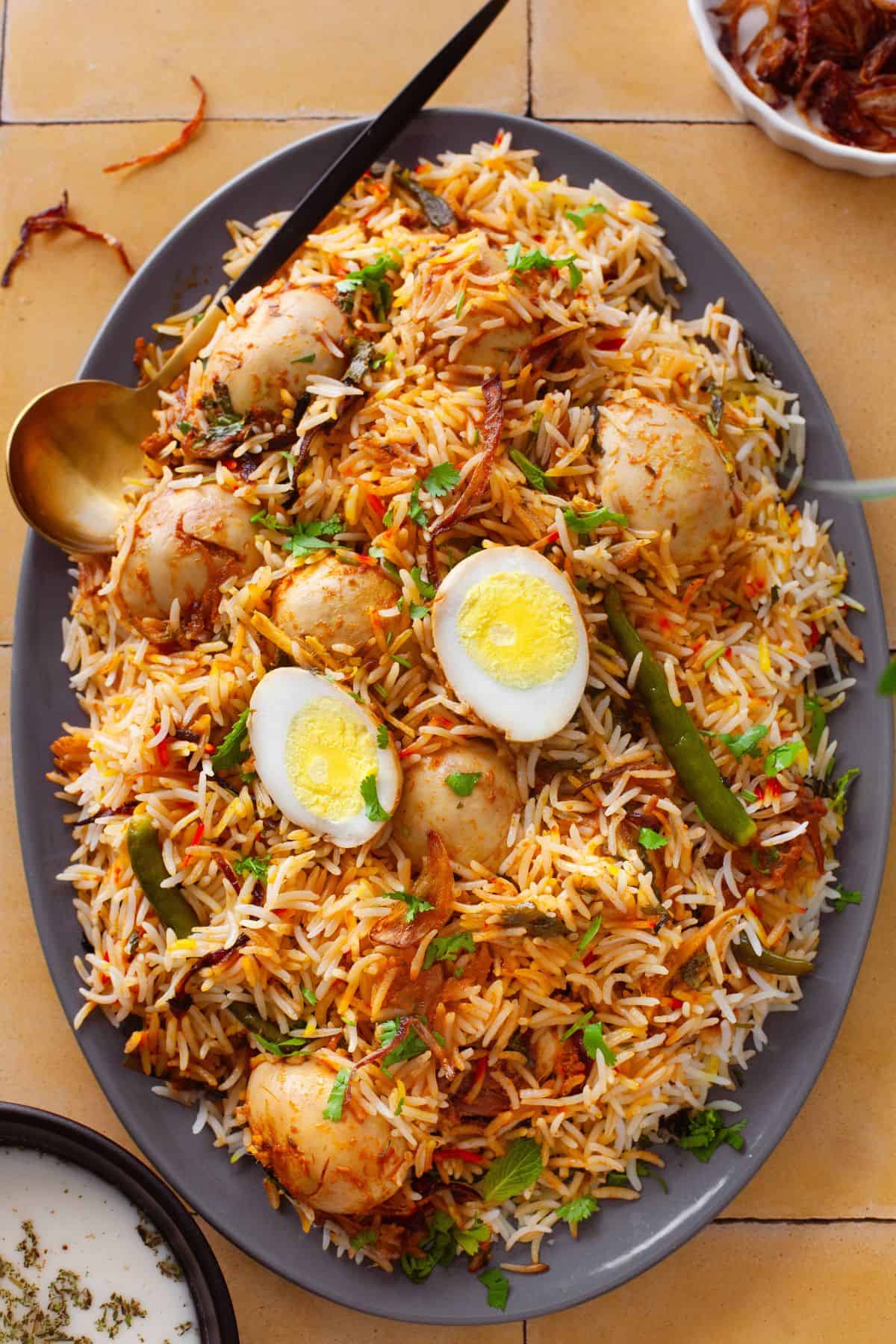 A platter of egg biryani garnished with boiled eggs, green chilies, fresh coriander, and crispy fried onions, served with a gold spoon on a tiled surface.