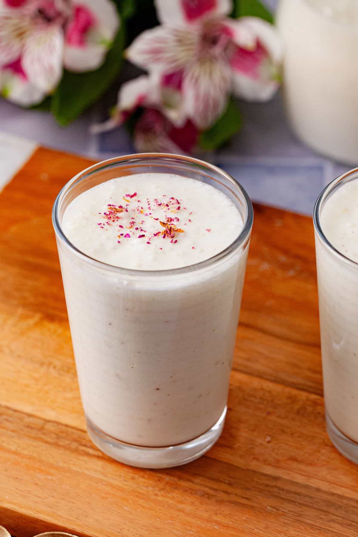 A glass of a creamy white lassi topped with a sprinkle of rose petals rests on a wooden surface.