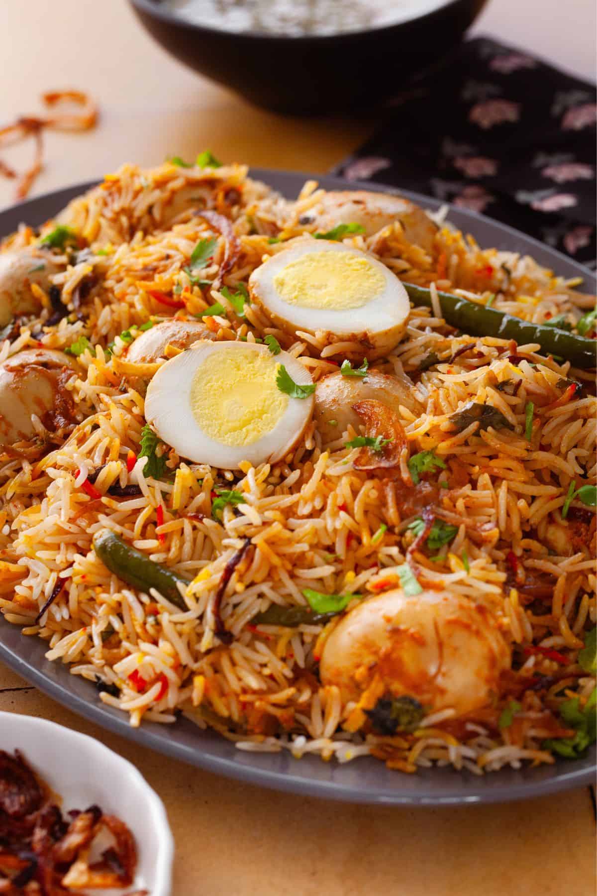 Side view of a plate of egg biryani garnished with sliced boiled eggs, green chilies, and fresh herbs, with long-grain rice mixed with spices.