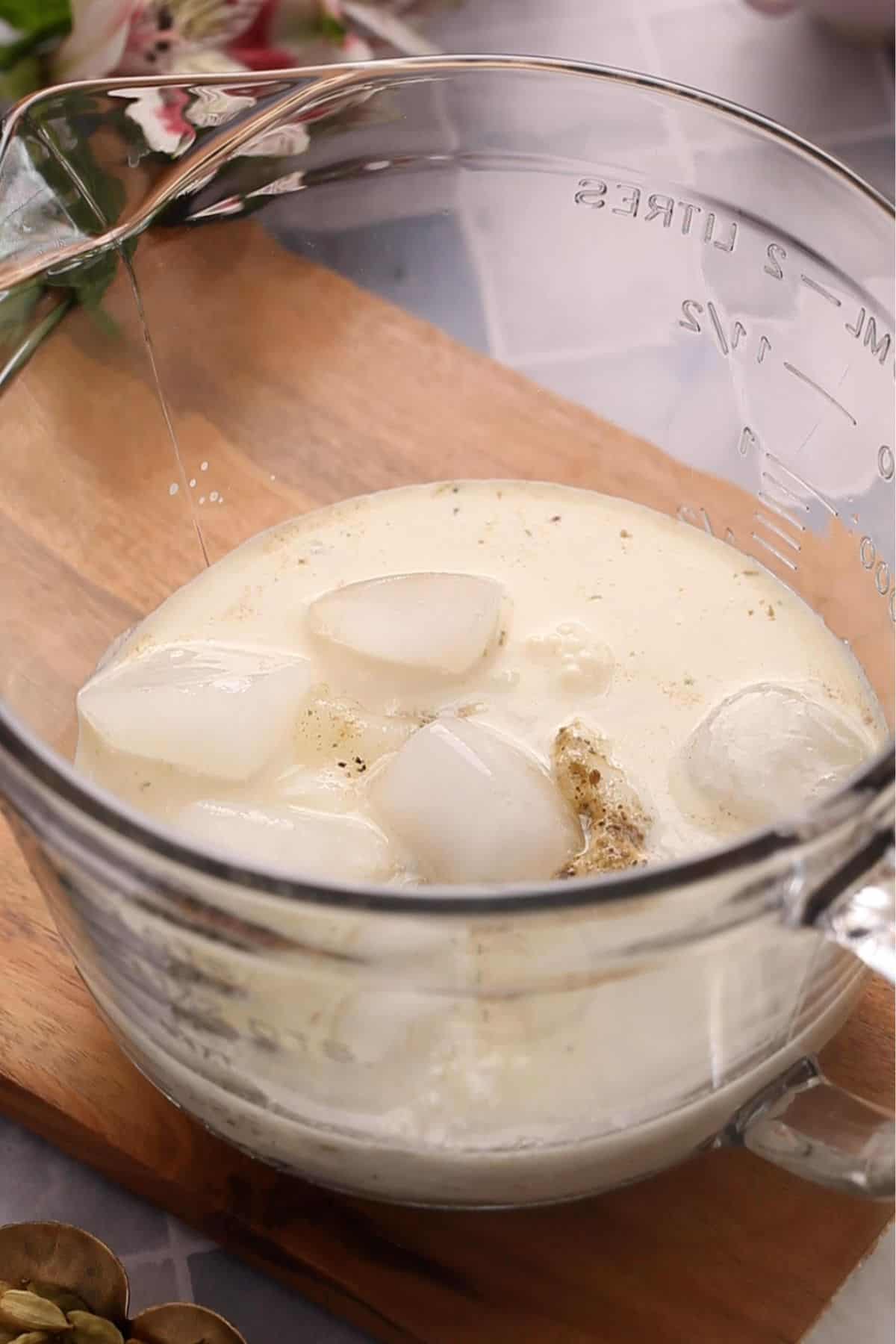 A clear glass jug on a wooden surface,with ingredients for making lassi.
