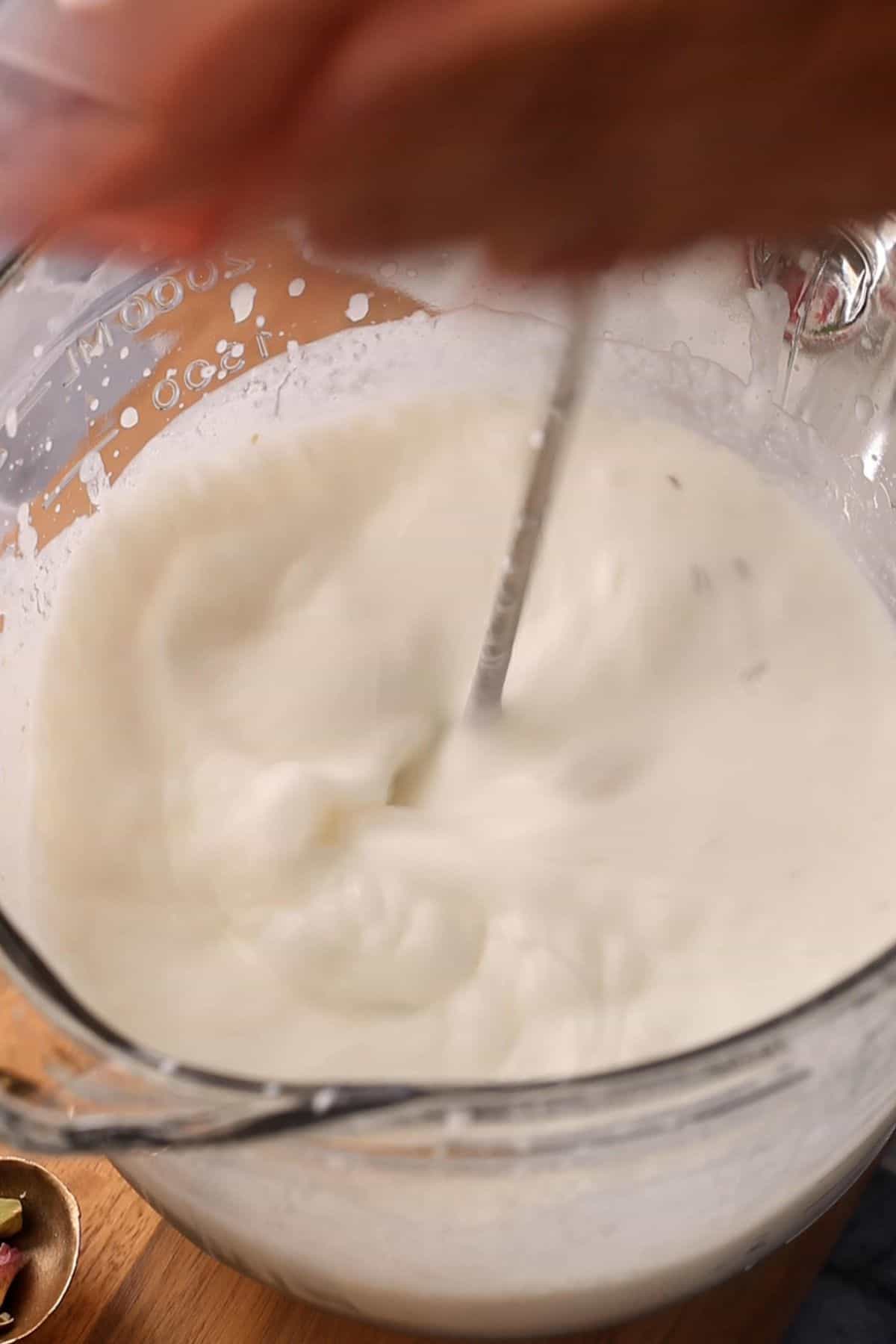 A hand whisk is used to beat yogurt mixture in a large glass cup placed on a wooden surface.