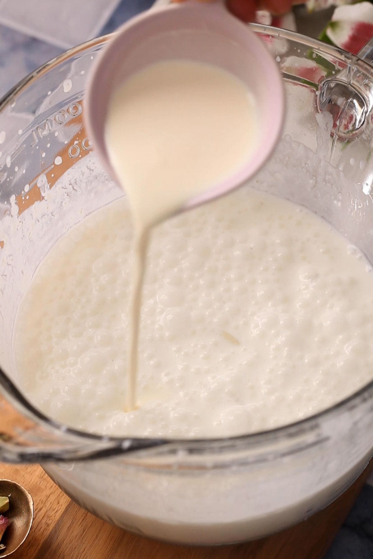 Pouring heavy cream from a small pink bowl into a larger glass bowl filled with frothy, yogurt mixture.