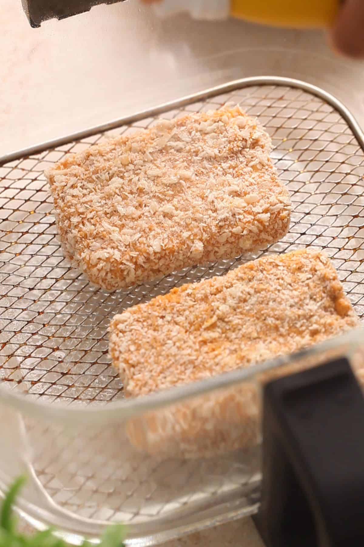 Two breaded rectangular pieces of paneer are placed on a wire rack inside an air fryer basket, ready to be cooked.
