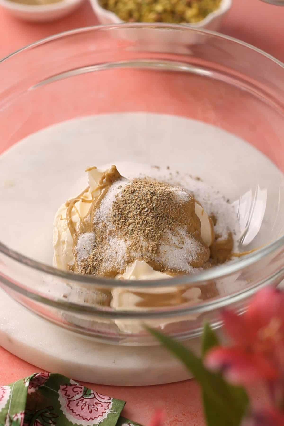 A glass bowl containing cream, pistachios, soft cheese  and spices sits on a marble surface.