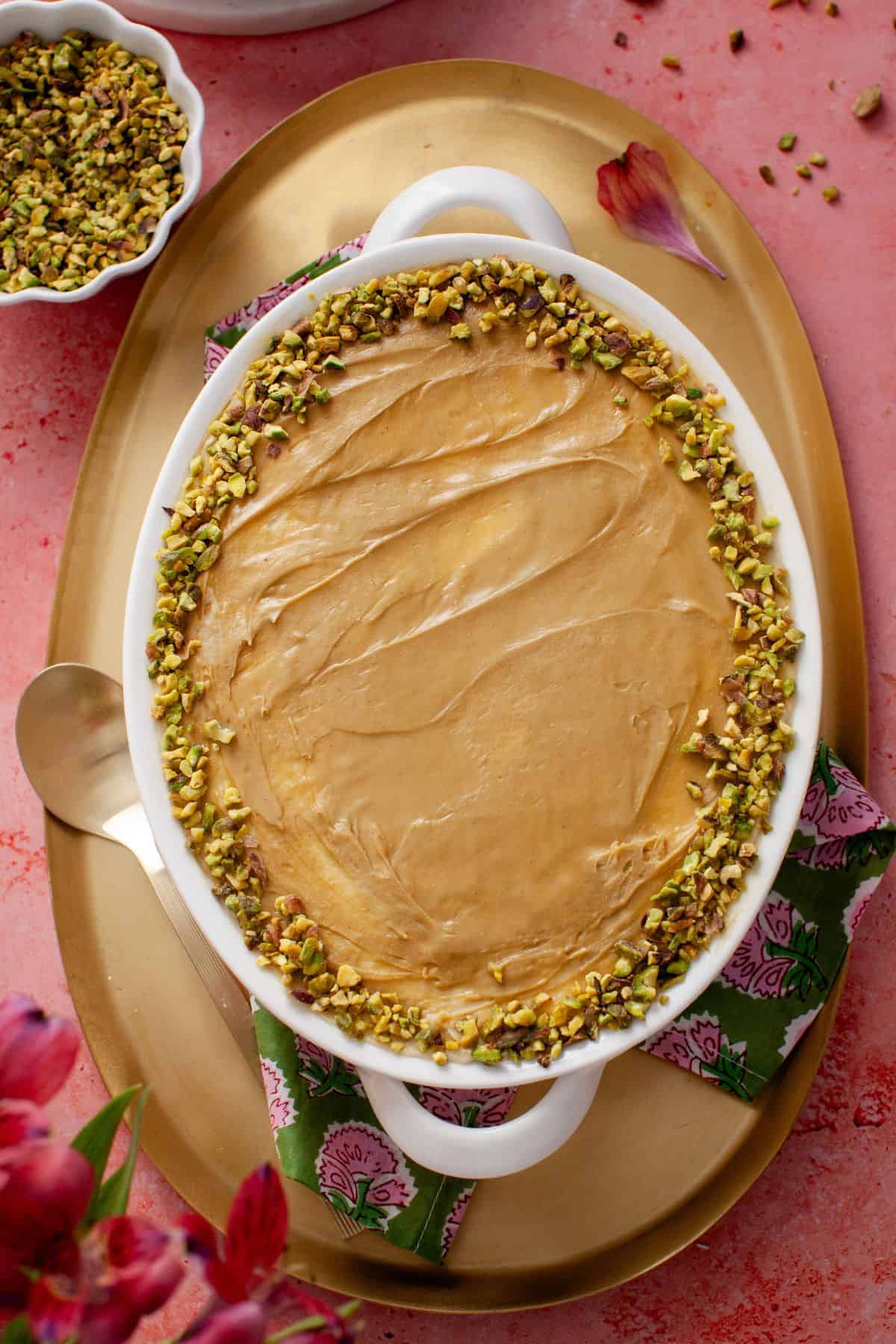 Oval dish filled with creamy pistachio tiramisu, topped with chopped pistachios around the edge, placed on a golden tray with a floral napkin.
