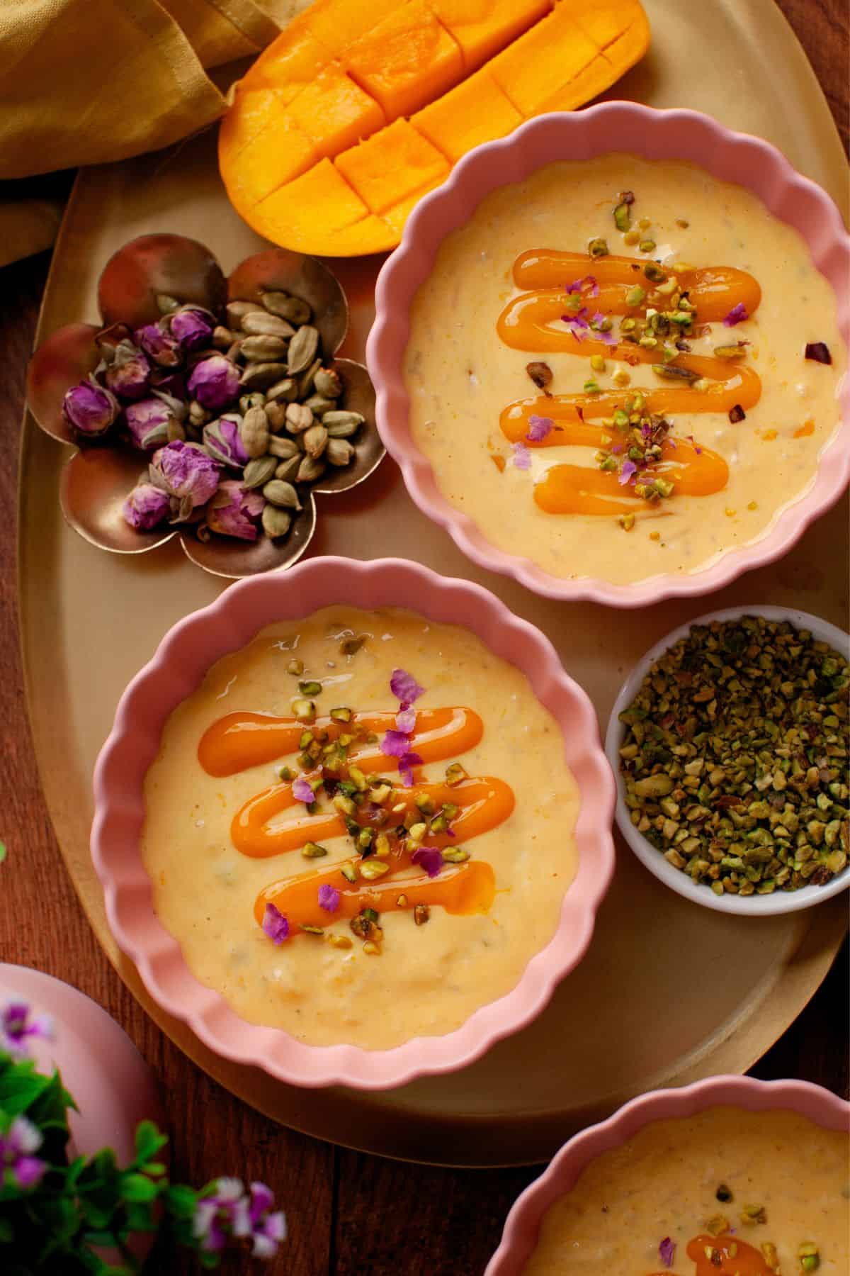 Three bowls of mango rice pudding topped with mango puree, chopped pistachios, and dried rose petals, are arranged on a tray with sliced mango, dried rosebuds, pistachios, and a bowl of chopped nuts.