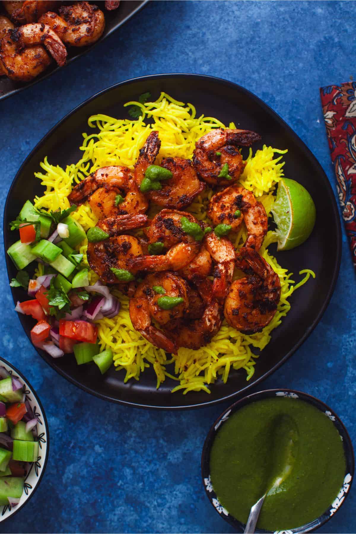 Tandoori shrimp served in a black plate over yellow rice with a drizzle of green chutney.