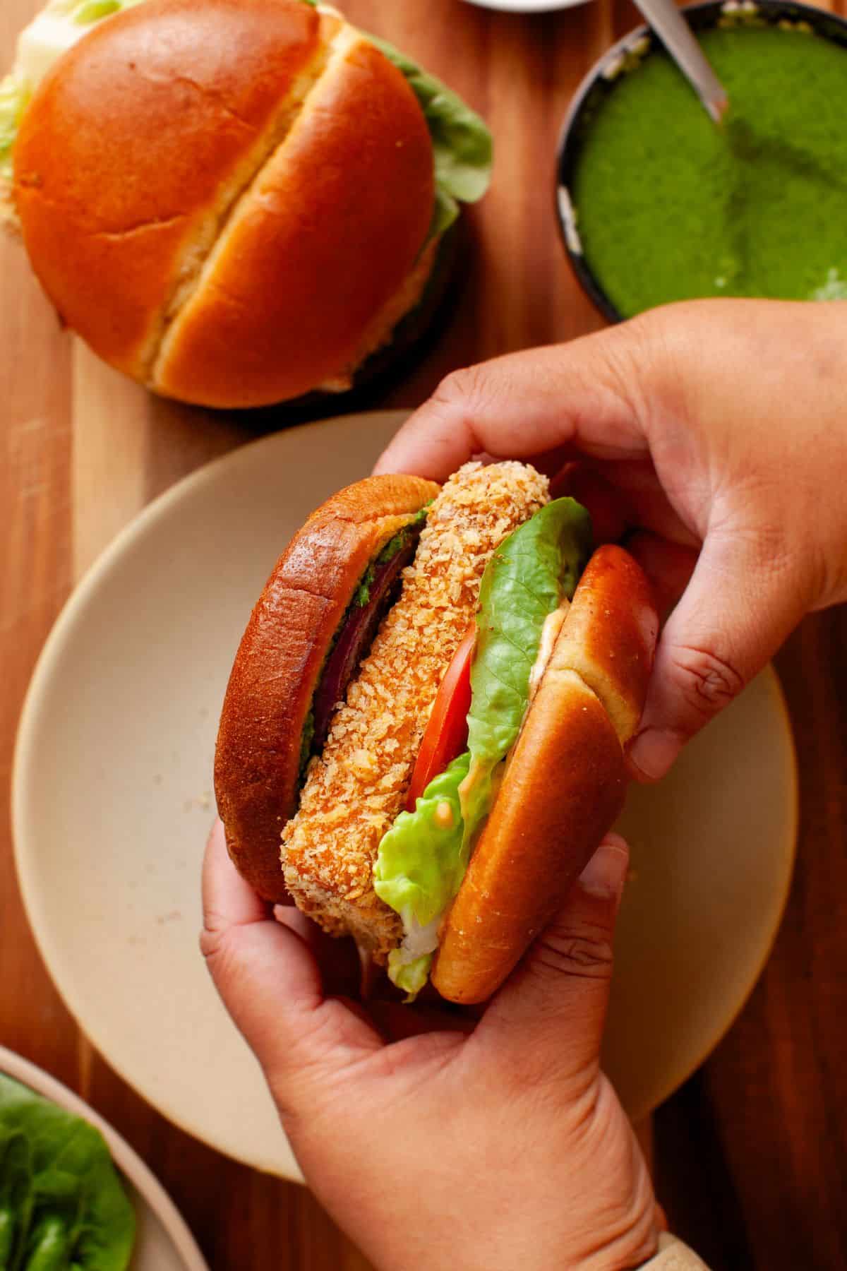 A person holds a burger with a breaded paneer patty, lettuce, tomato, and a brioche bun over a plate.