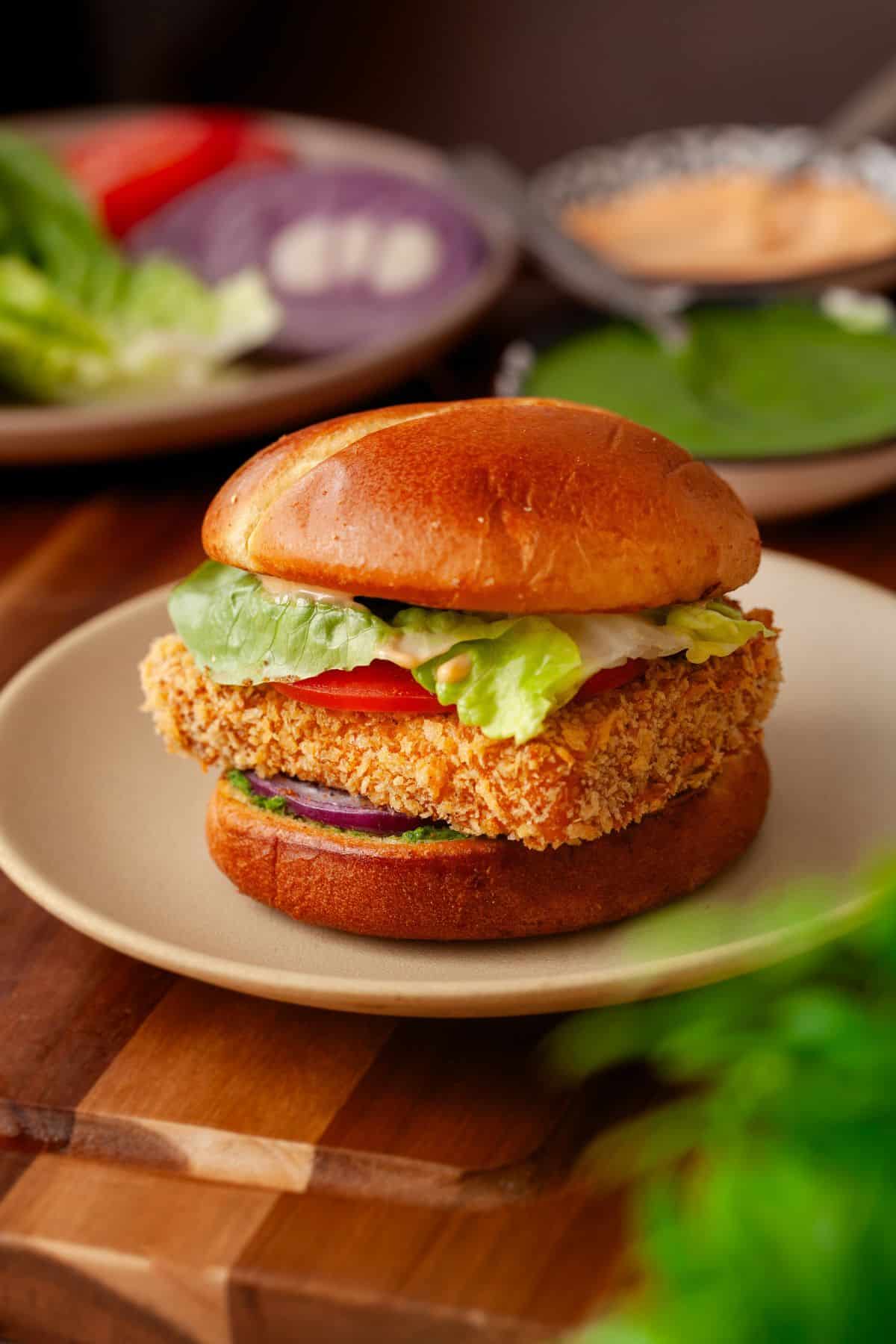 A crispy breaded paneer burger with lettuce, tomato, and sauce on a toasted bun, served on a plate with sliced vegetables and sauce in the background.