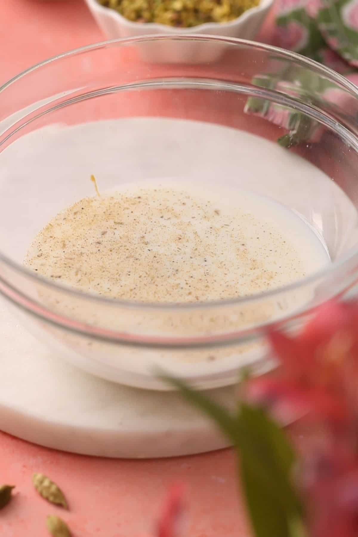 A glass bowl containing milk and ground spices sits on a white marble surface, with a blurry pink flower and green cardamom pods in the foreground.