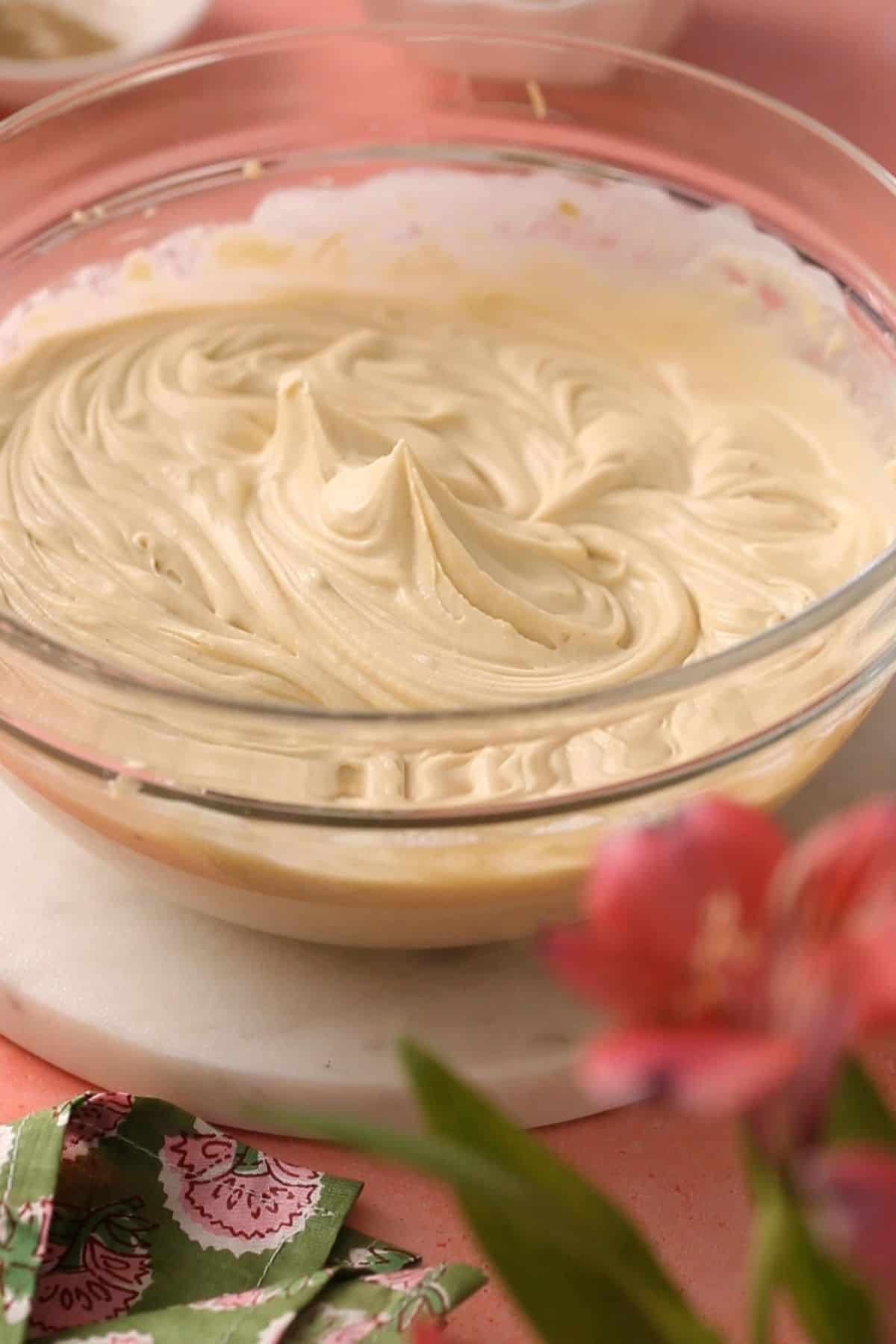 A glass bowl filled with creamy tiramisu cream sits on a marble surface, with a pink flower and green patterned napkin in the foreground.