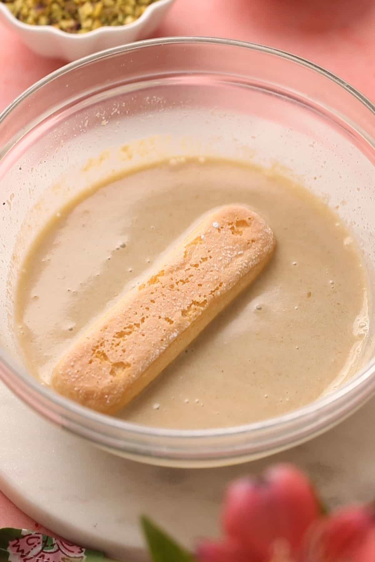 A ladyfinger biscuit partially soaked in a bowl of creamy pistachio milk.