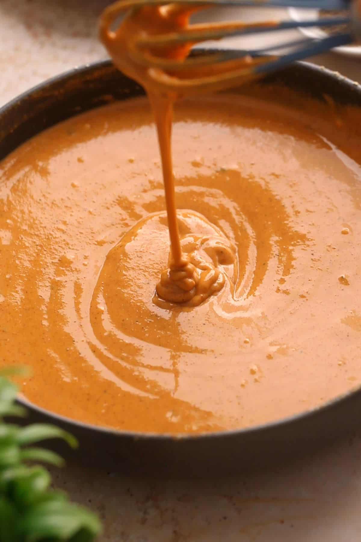 A whisk drizzles pakora batter into a bowl filled with the same sauce, creating swirls on the surface.