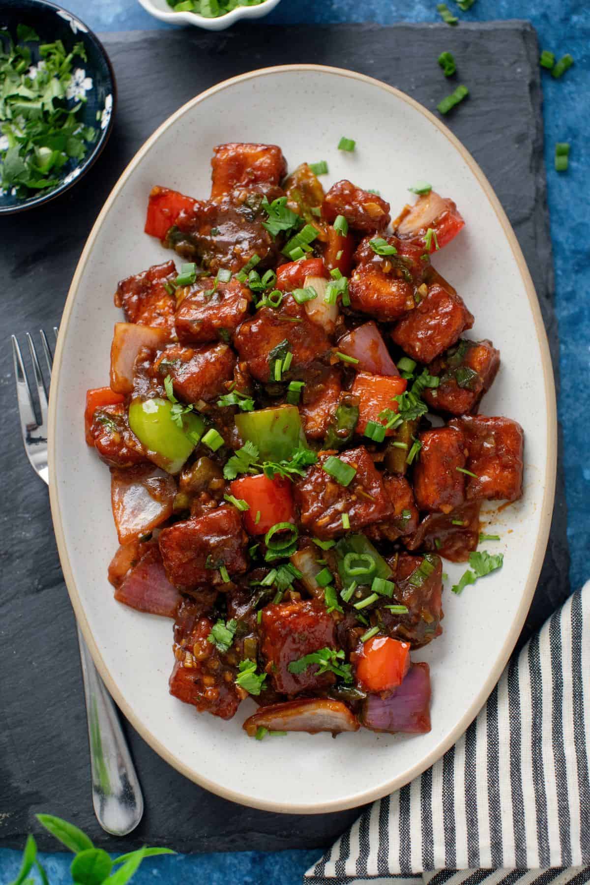 Chilli Paneer served on an oval platter placed on a blue background.