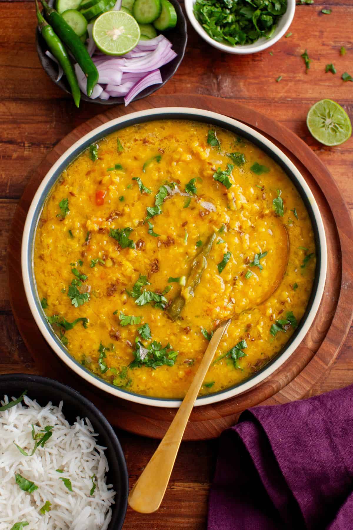 A bowl of masoor dal garnished with fresh cilantro, with a spoon inside, surrounded by a bowl of white rice, a plate of sliced cucumber, onions, green chilies, and a small bowl of chopped herbs on a wooden table.