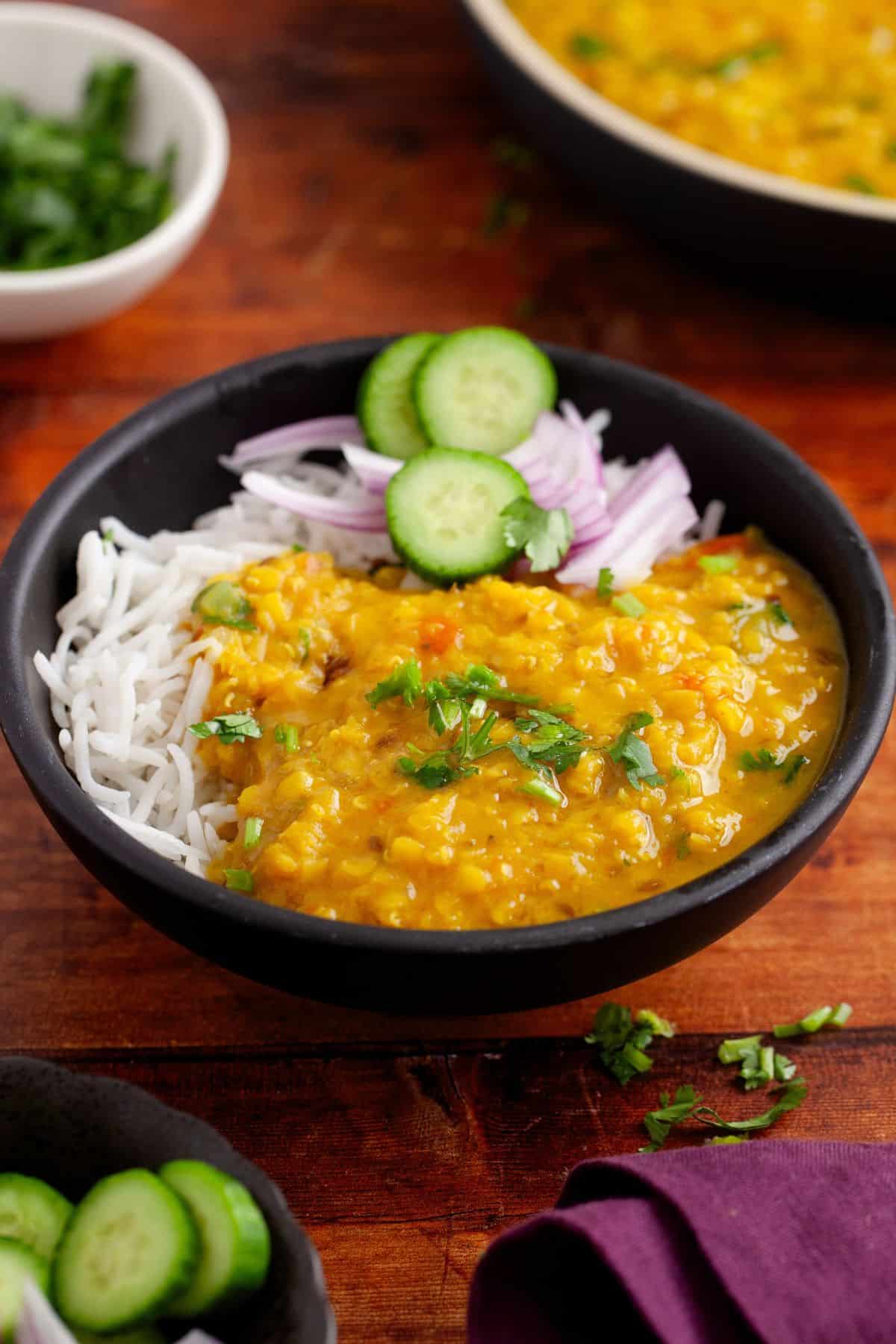A black bowl of red lentils dal served with white rice, topped with sliced cucumbers, red onions, and fresh cilantro, placed on a wooden table.