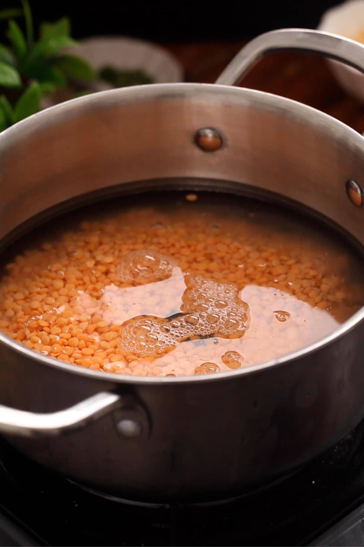 A stainless steel pot filled with red lentils soaking in water sits on a stovetop. Green herbs and a bowl are blurry in the background.