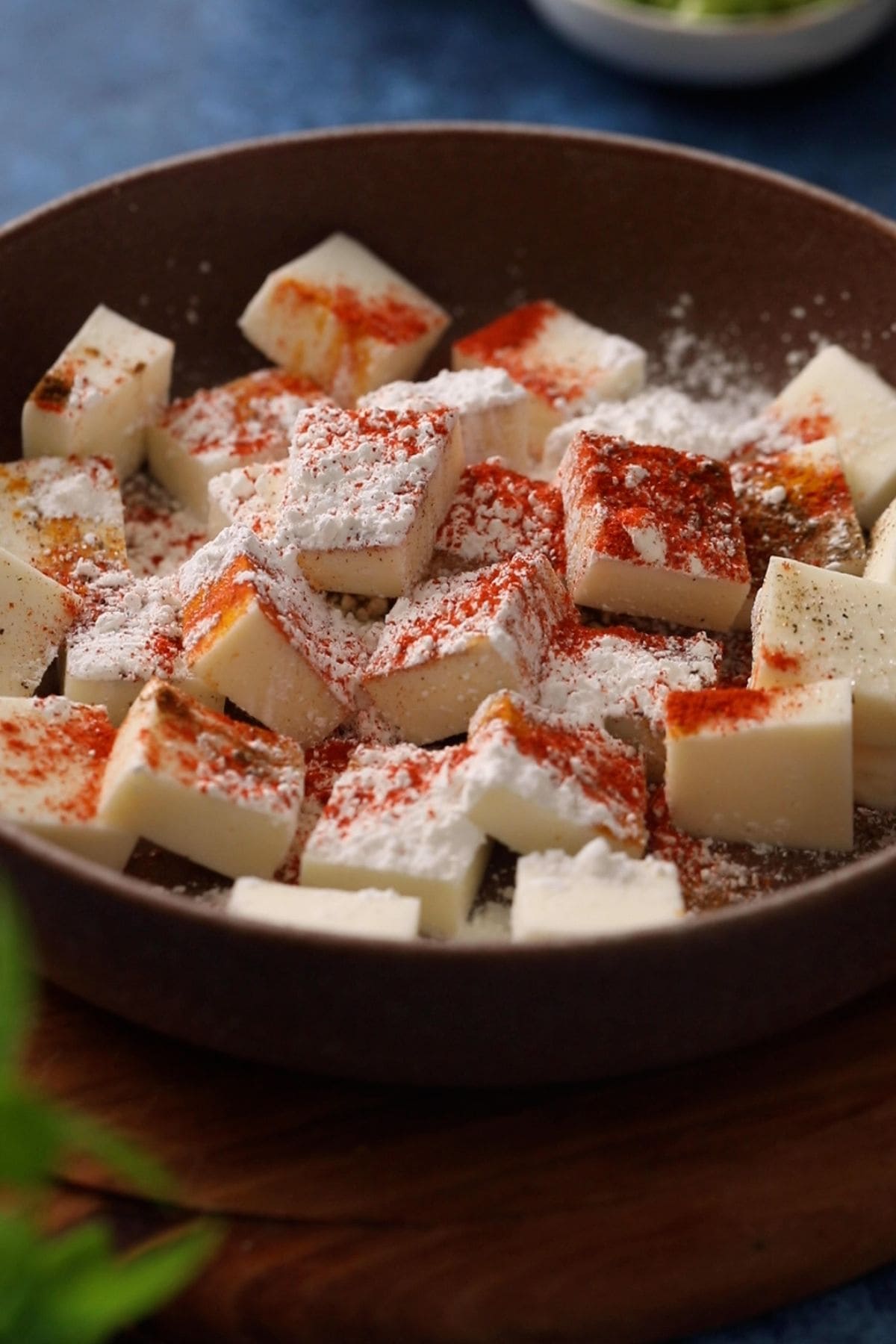 Spices and cornstarch is sprinkled on paneer cubes placed in a bowl.