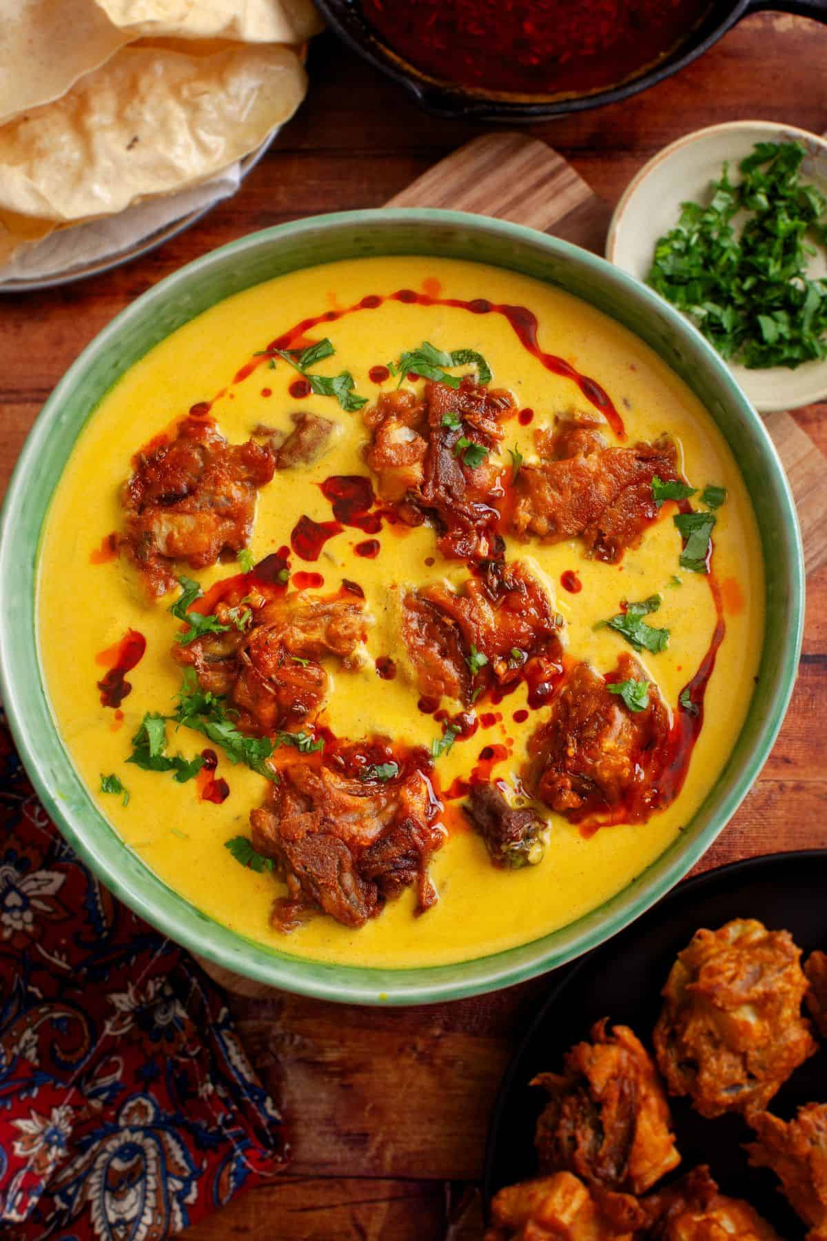 A green bowl filled with kadhi topped with fried pakora, garnished with chopped cilantro and ghee tadka. A plate of herbs and a dish of snacks are visible in the background.