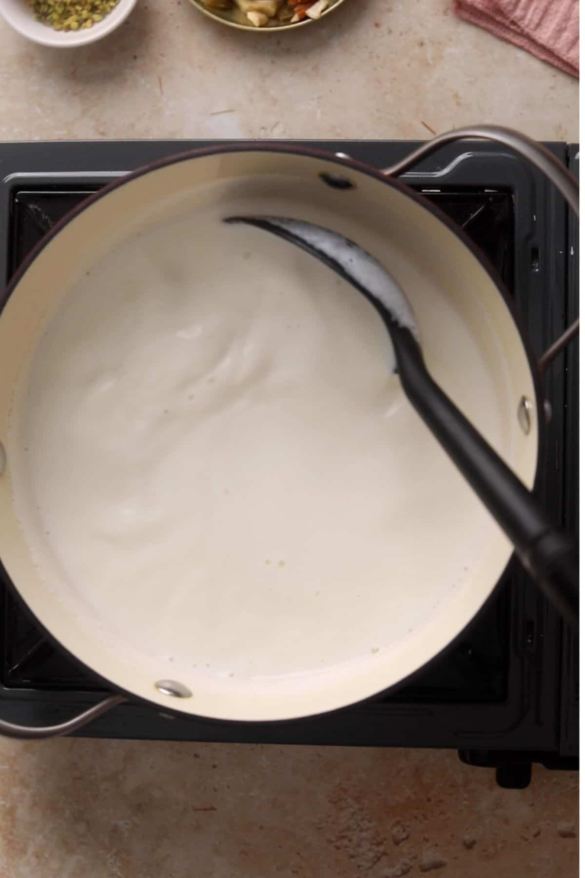 A saucepan filled with milk is being stirred with a black ladle on a stovetop.