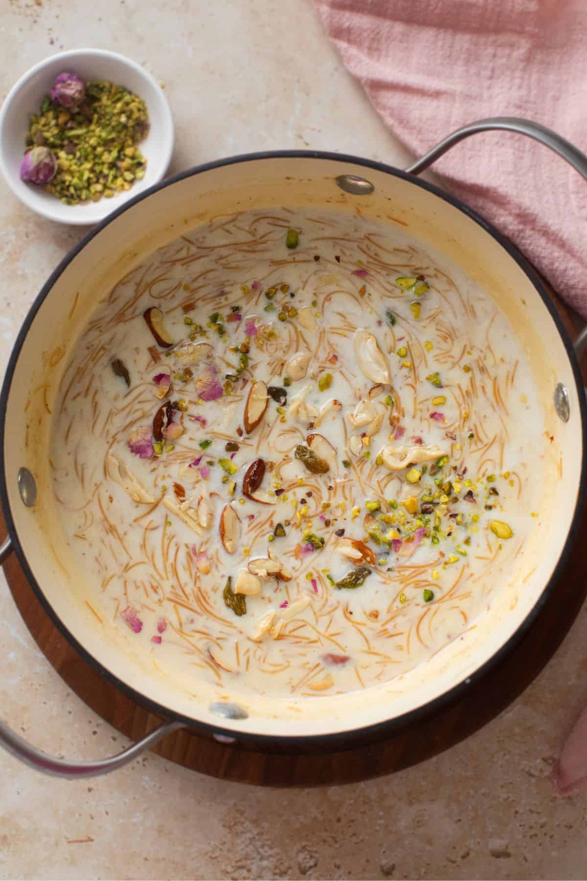 A pot of creamy vermicelli dessert garnished with chopped pistachios and rose petals. A small bowl with additional pistachios and rosebuds is nearby on a light surface, next to a pink cloth.