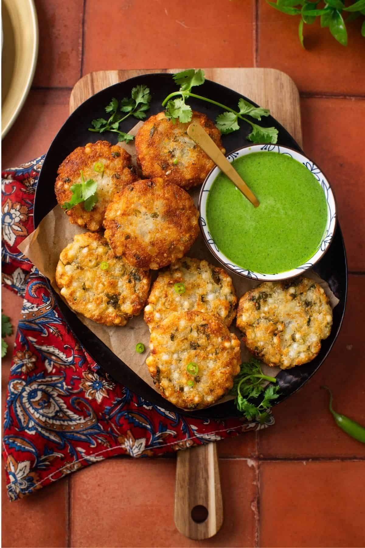 A black platter of deep fried and air fried sabudana vada served with green chutney and garnished with cilantro leaves and green chillies. 