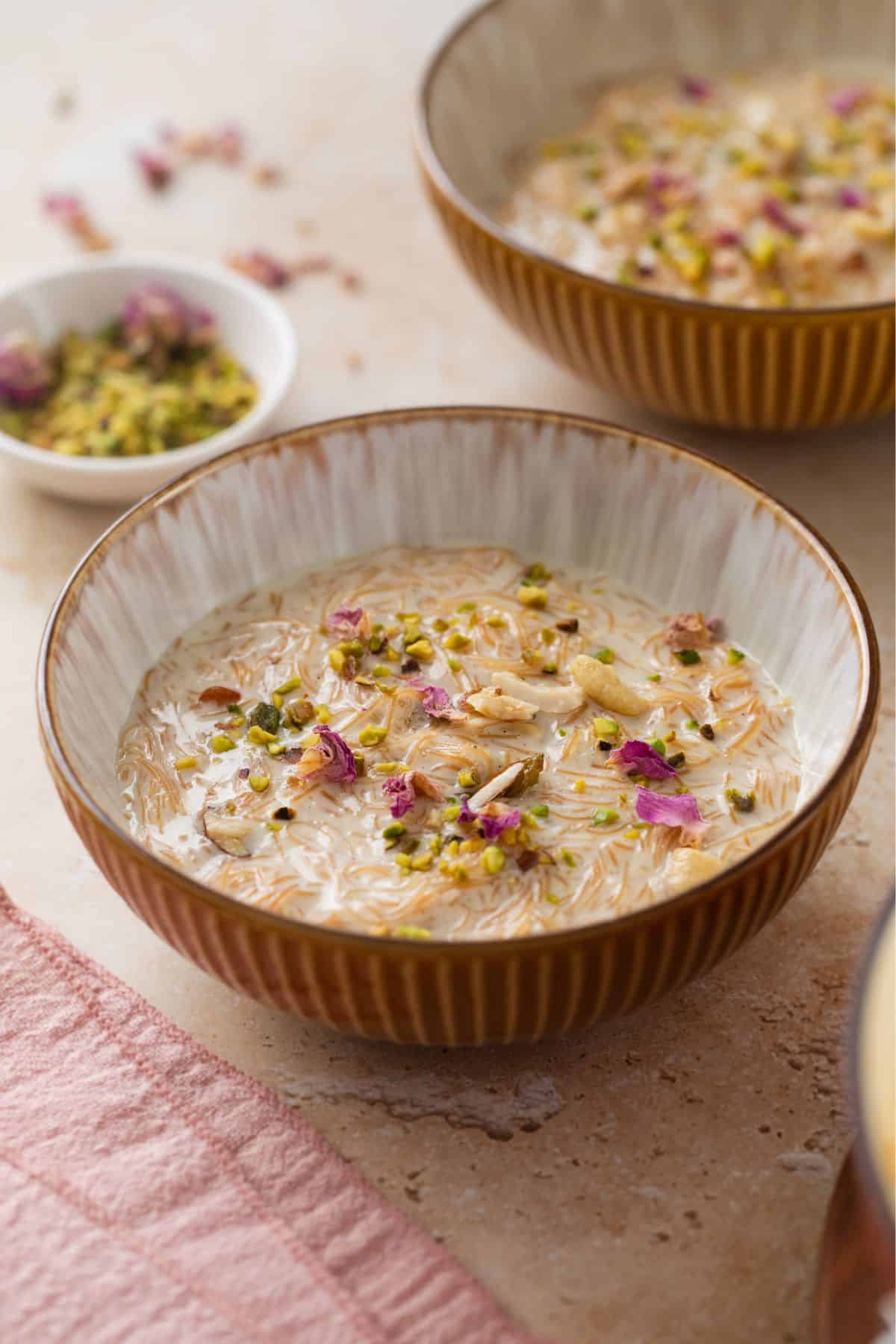 Side image of a bowl of A bowl of creamy seviyan kheer garnished with chopped nuts and dried rose petals, placed on a light surface with a pink cloth.