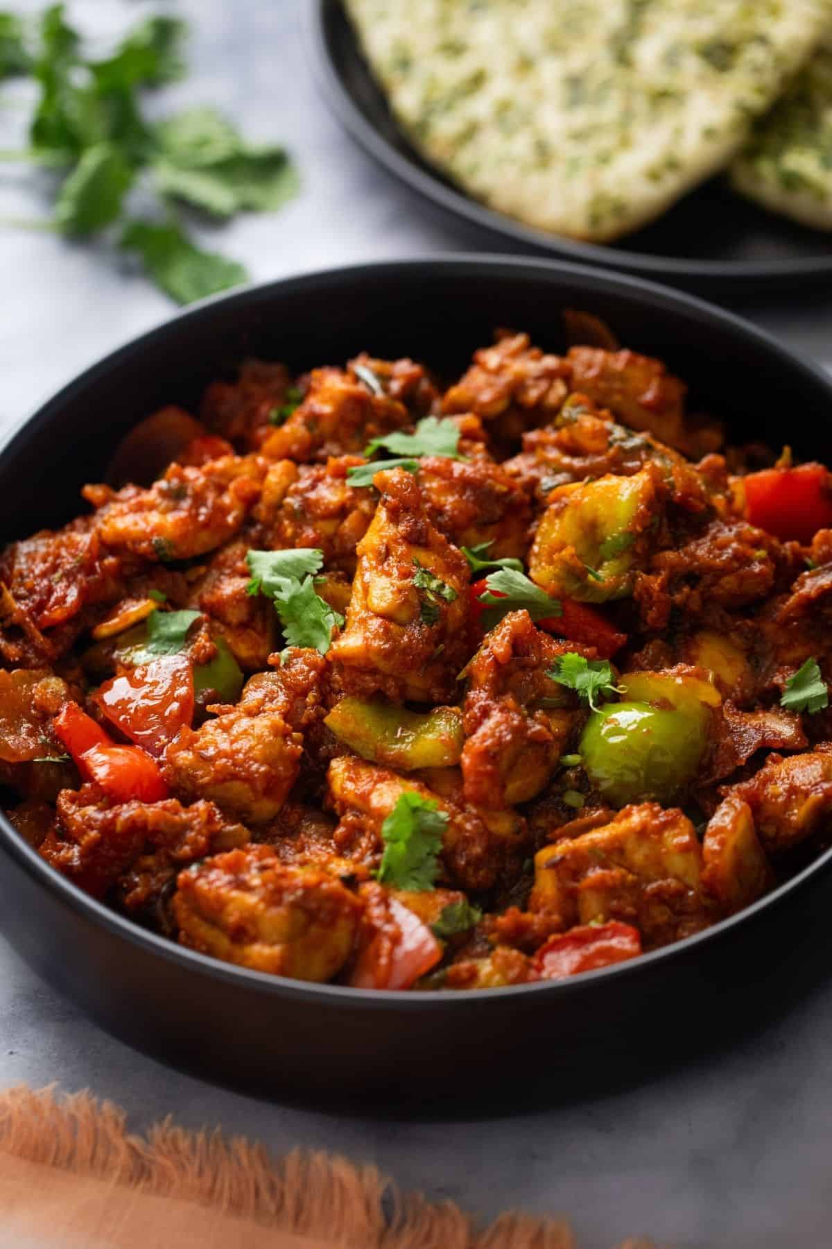 Chicken jalfrezi in a black bowl with naan in background.