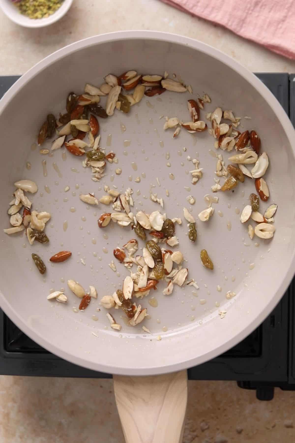 A ceramic pan on a stove contains sautéed nuts and raisins in oil, including cashews, almonds, and golden raisins.