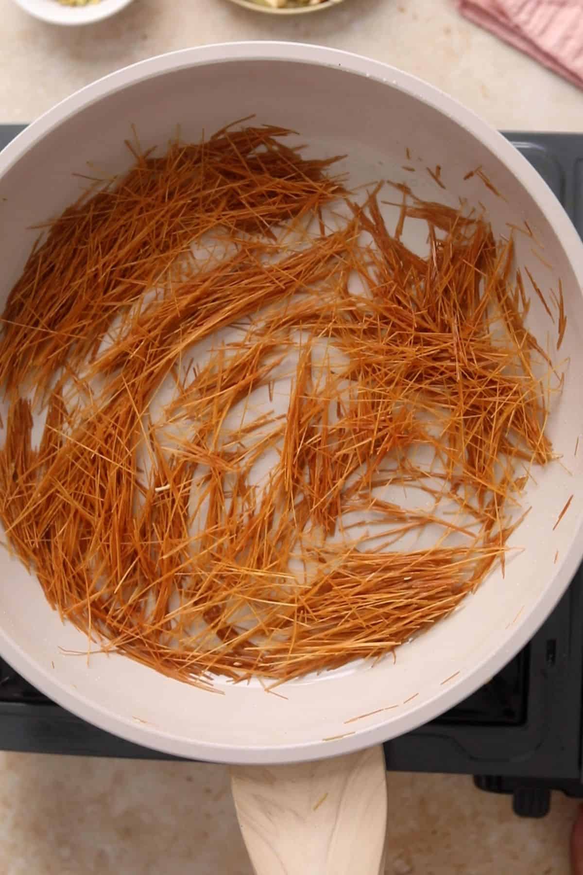 A white pan on a stove contains thin, golden-brown toasted seviyan, with a wooden spatula resting on the edge of the pan.