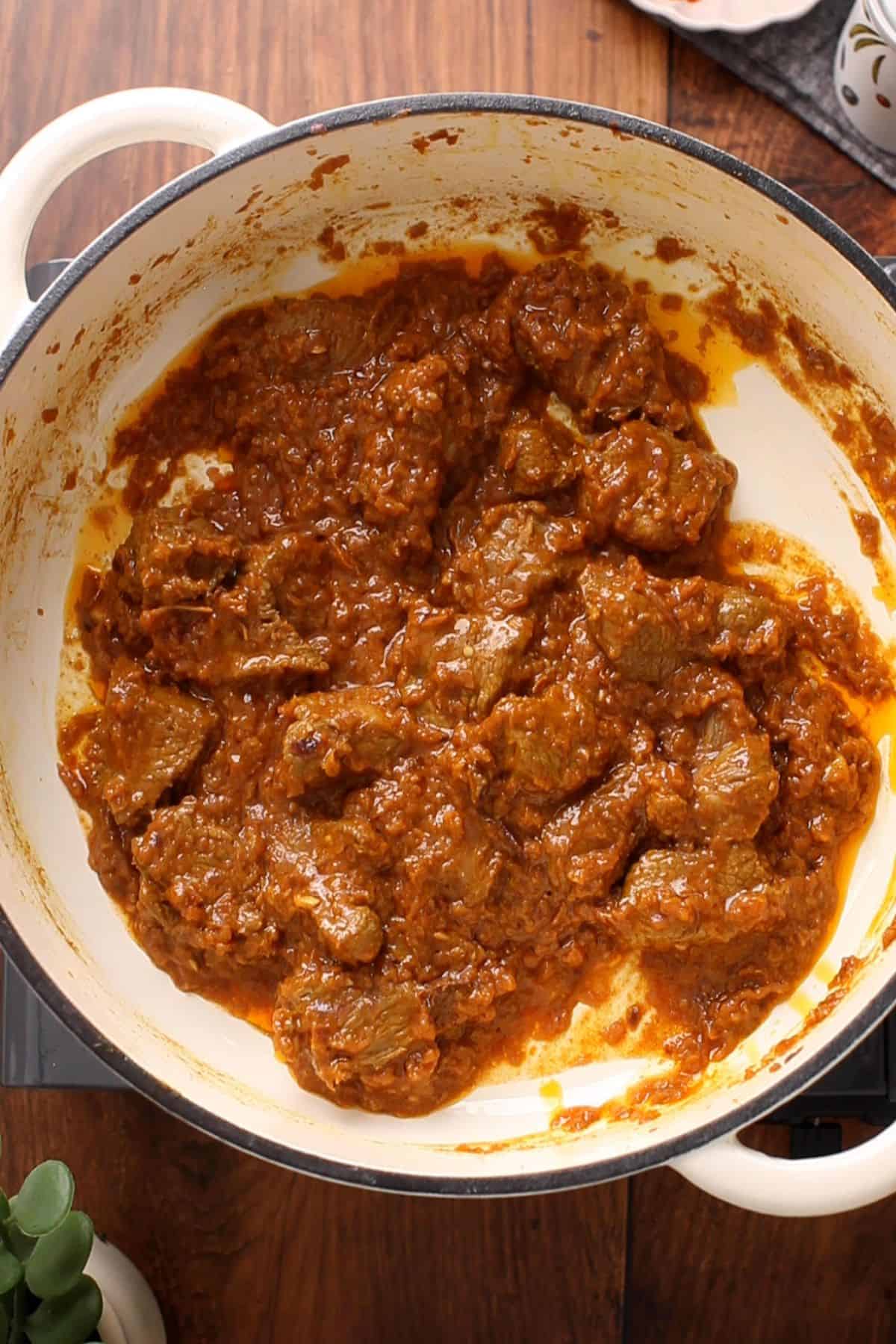 Lamb is cooking with onion tomato masala in a white pot.