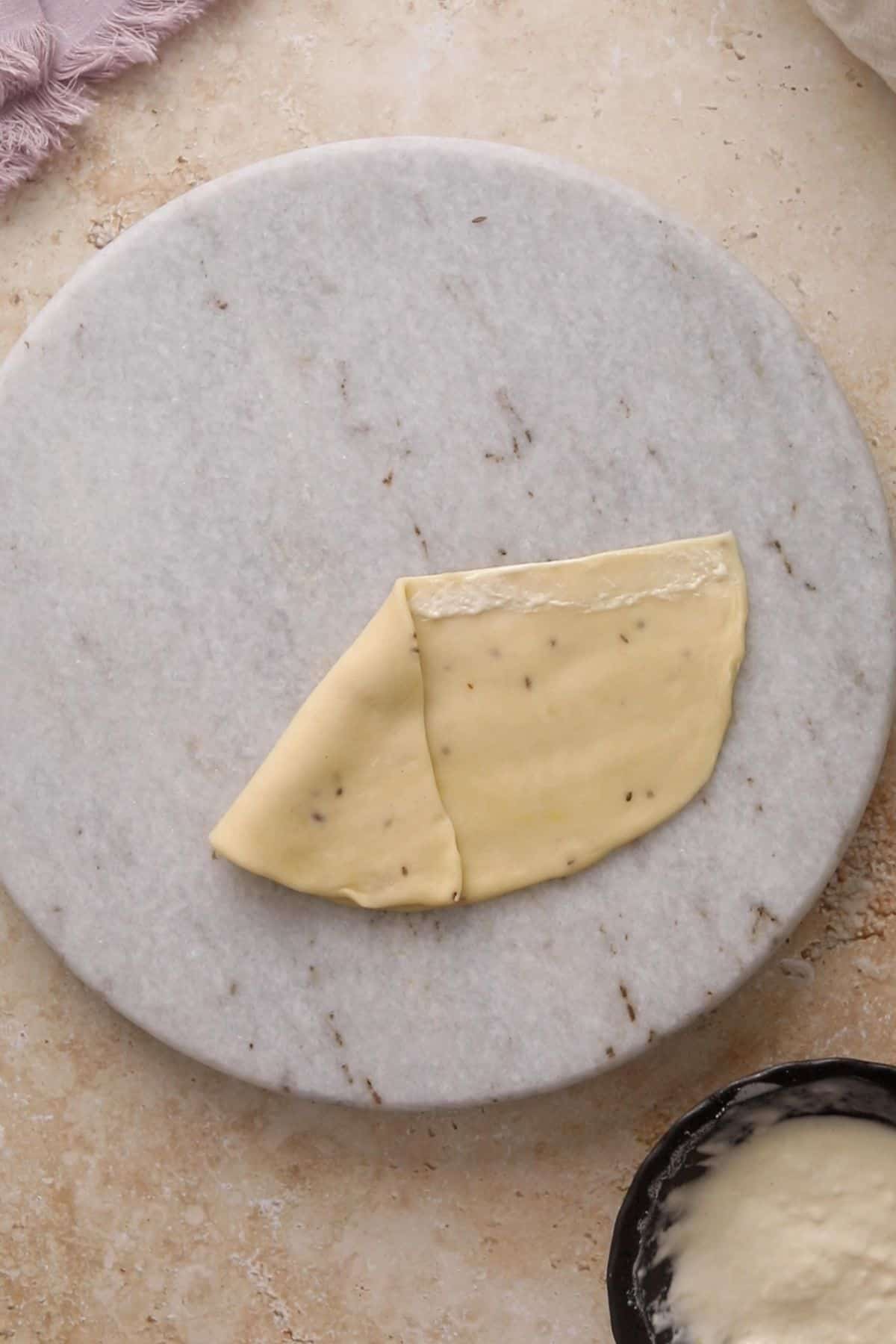 A partially folded piece of dough with herbs rests on a round marble board atop a beige surface, with a bowl of flour and a pink cloth visible at the edges.