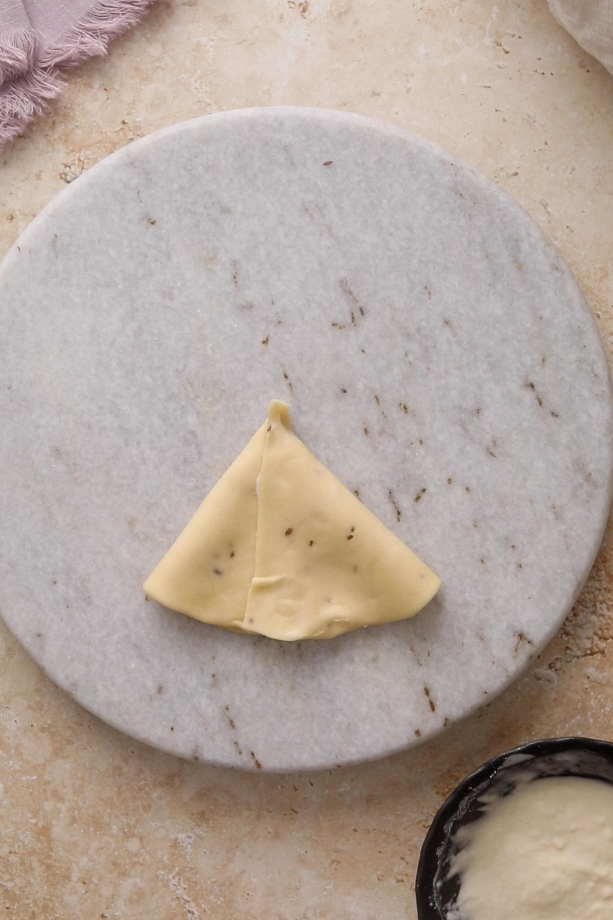 A single, triangular piece of dough is folded on a round marble board.
