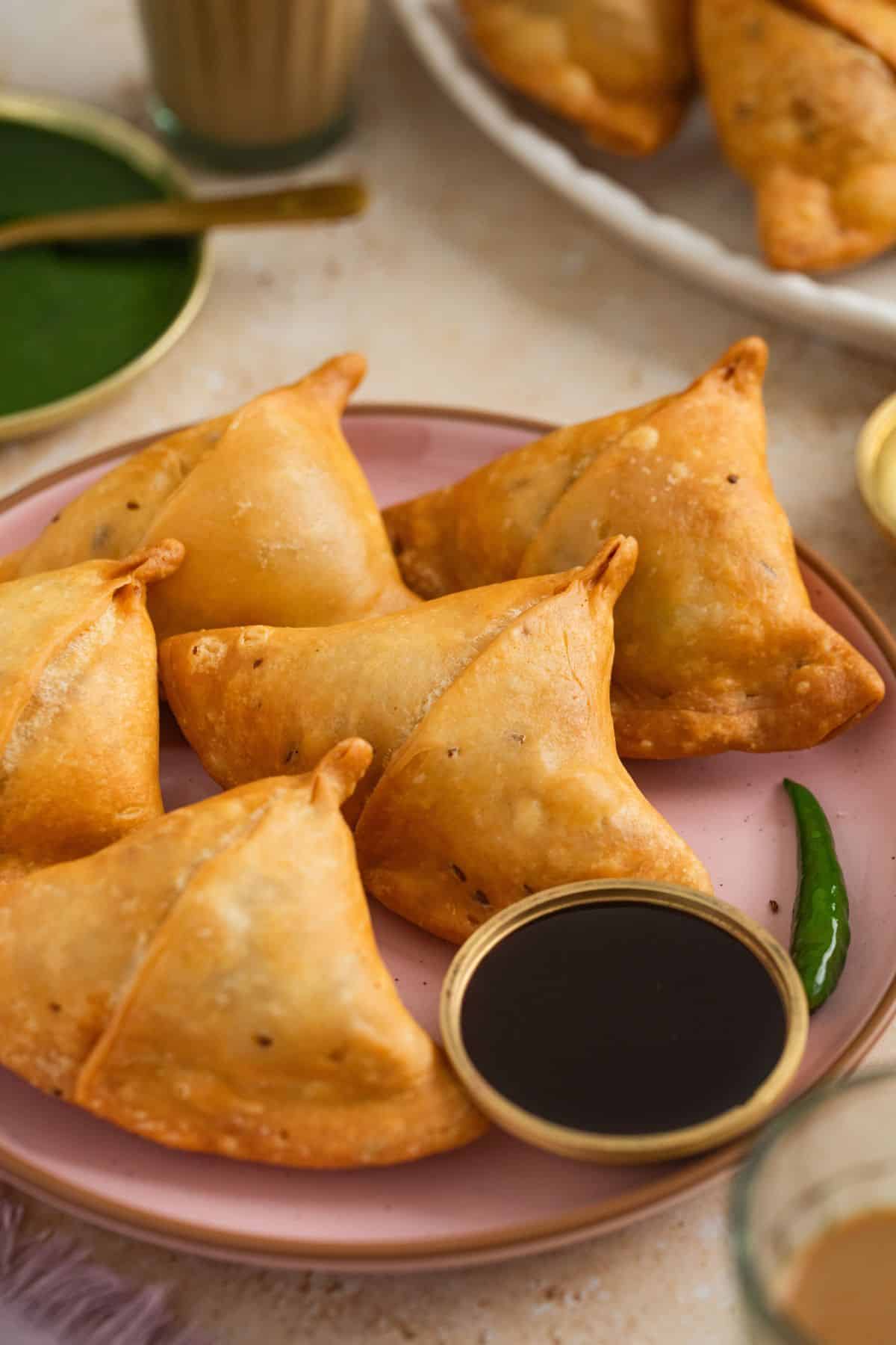 A pink plate with five golden-brown samosas, served with a small bowl of tamarind and green chutney.Other dishes and a glass of chai are visible in the background.