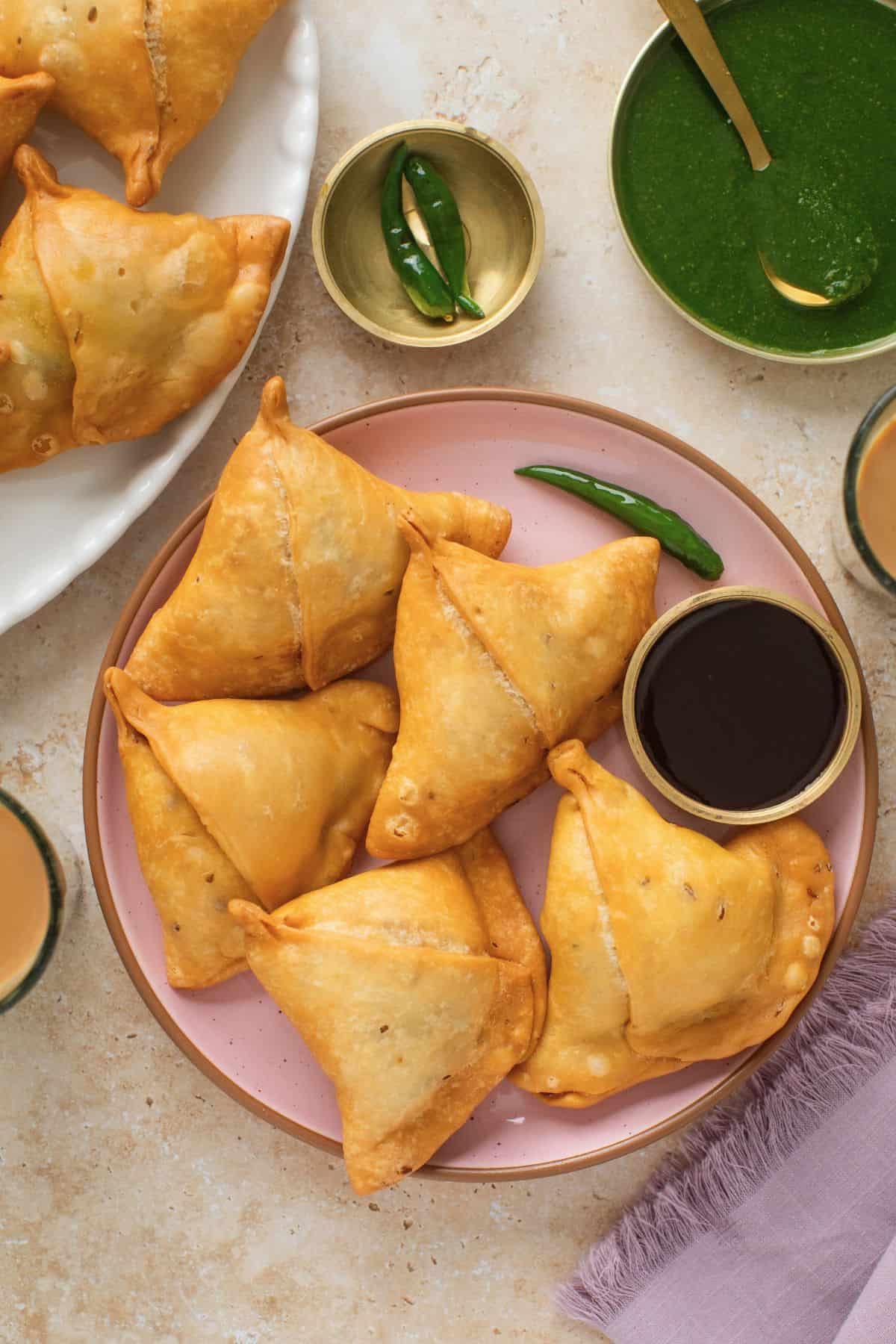 Top view of samosas on a pink plate with friend green chillies abd chutneys.