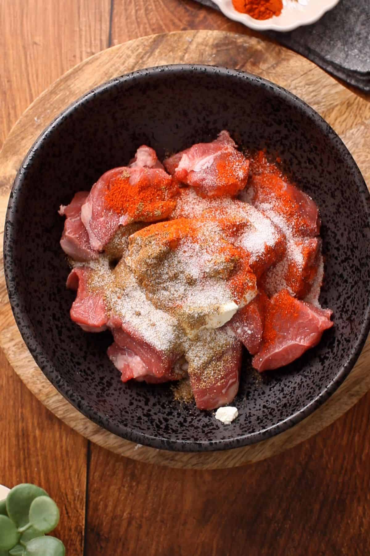 Raw lamb meat and marinade ingredients are added to a large black bowl.