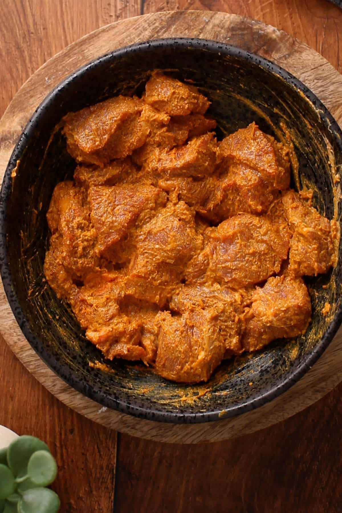 Lamb meat is coated in yogurt and spices in a black bowl placed on wooden surface.
