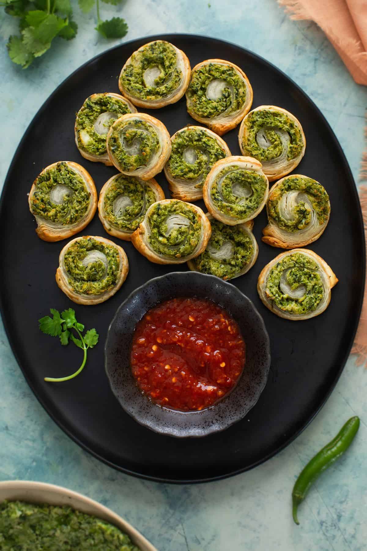 Hara Bhara Kebab pinwheels placed on a large black platter with chilli garlic sauce.