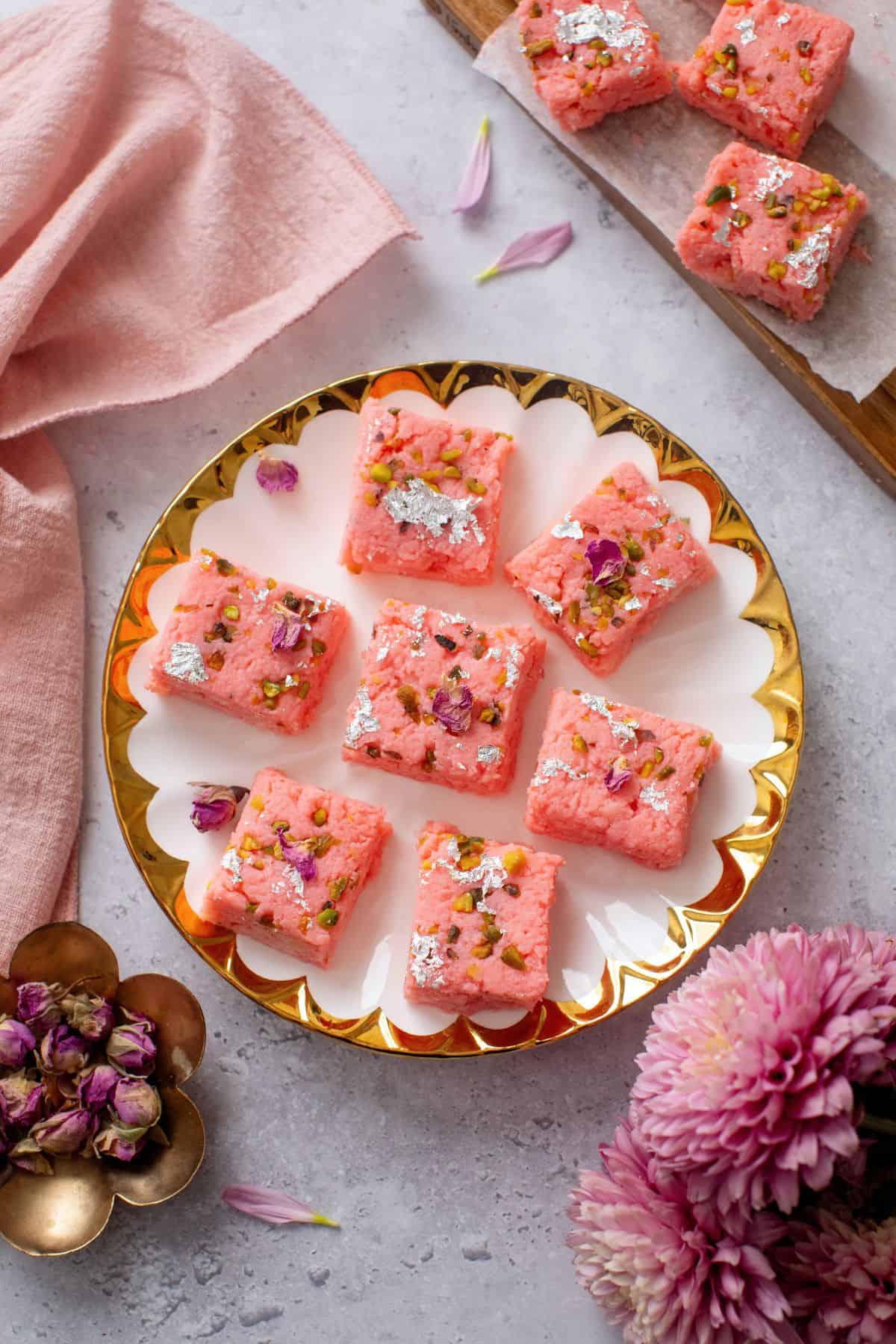 Pink Paneer kalakand placed on a white and gold rimmed plate with purpse flowers and dried roses. 