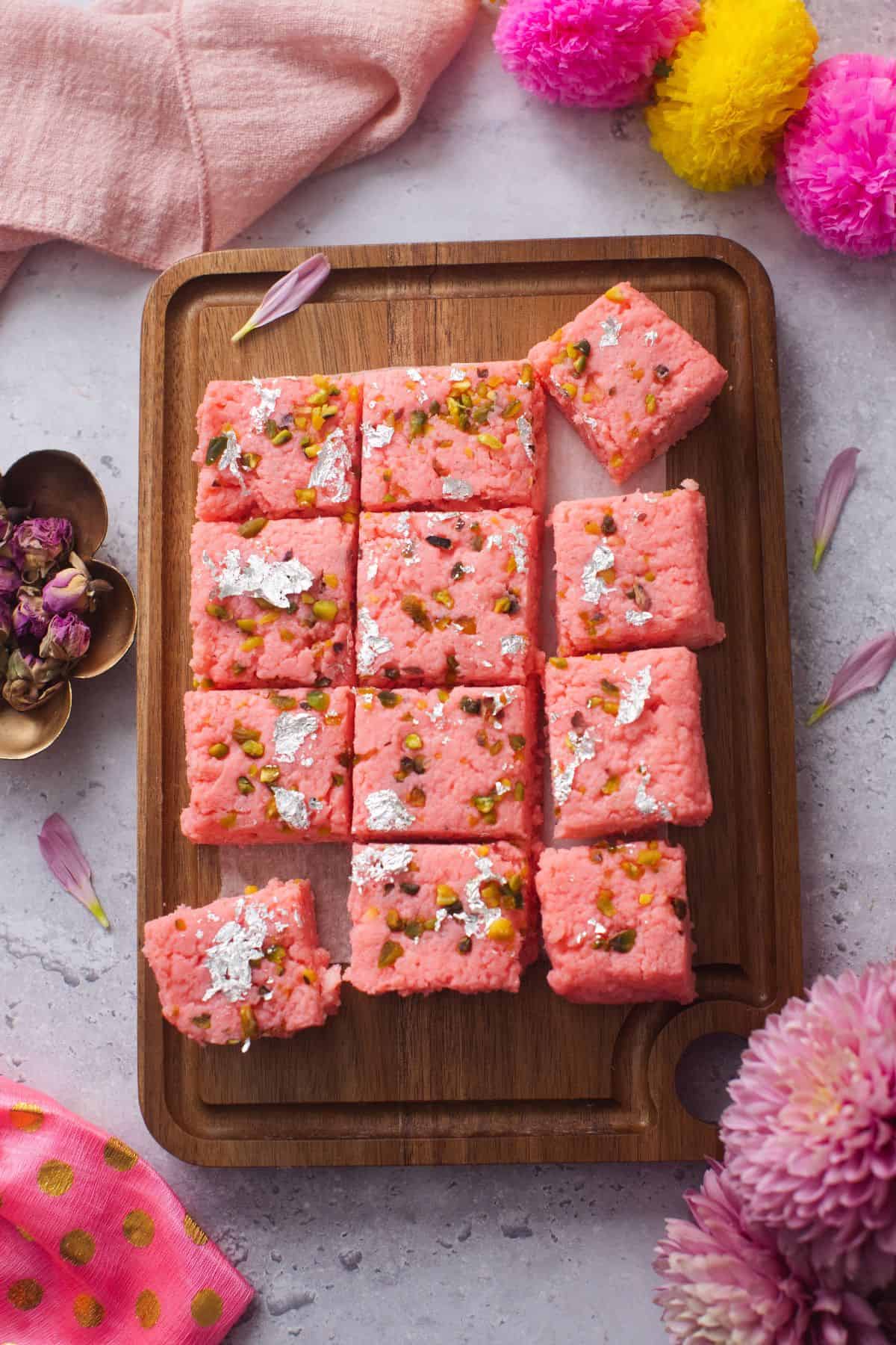 Cut up kalakand is place on a brown wooden board with pink and yellow marigold garland.