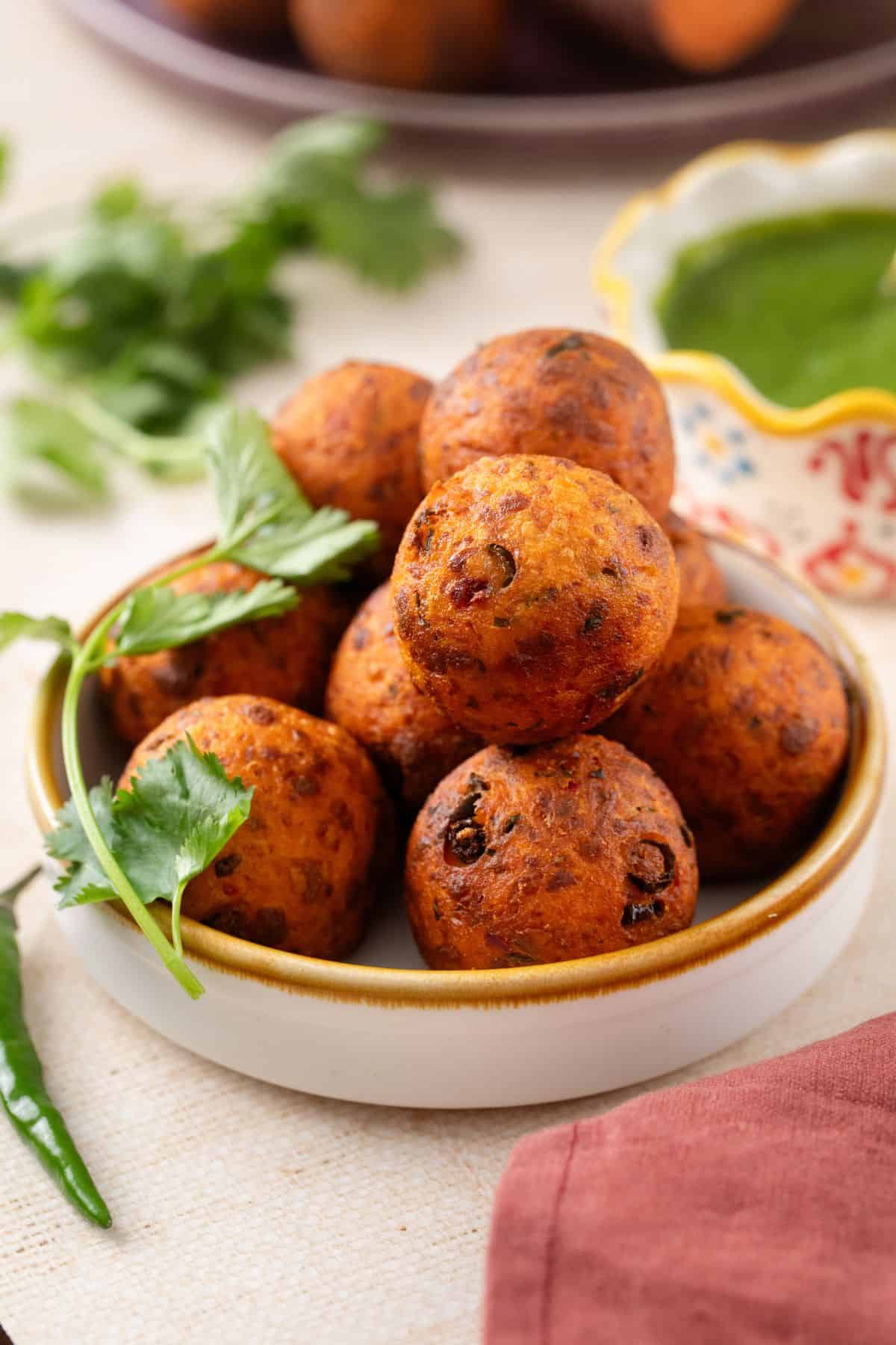 Sweet potato kofta stacked in a white bowl with brown rim. there's a bowl of green chutney at the back. 