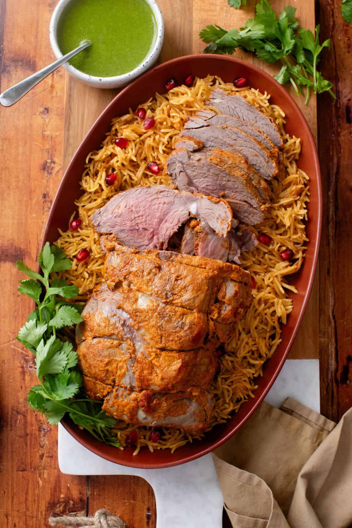 Roasted lamb raan rests on a bed of basmati rice with a bowl of green chutney.
