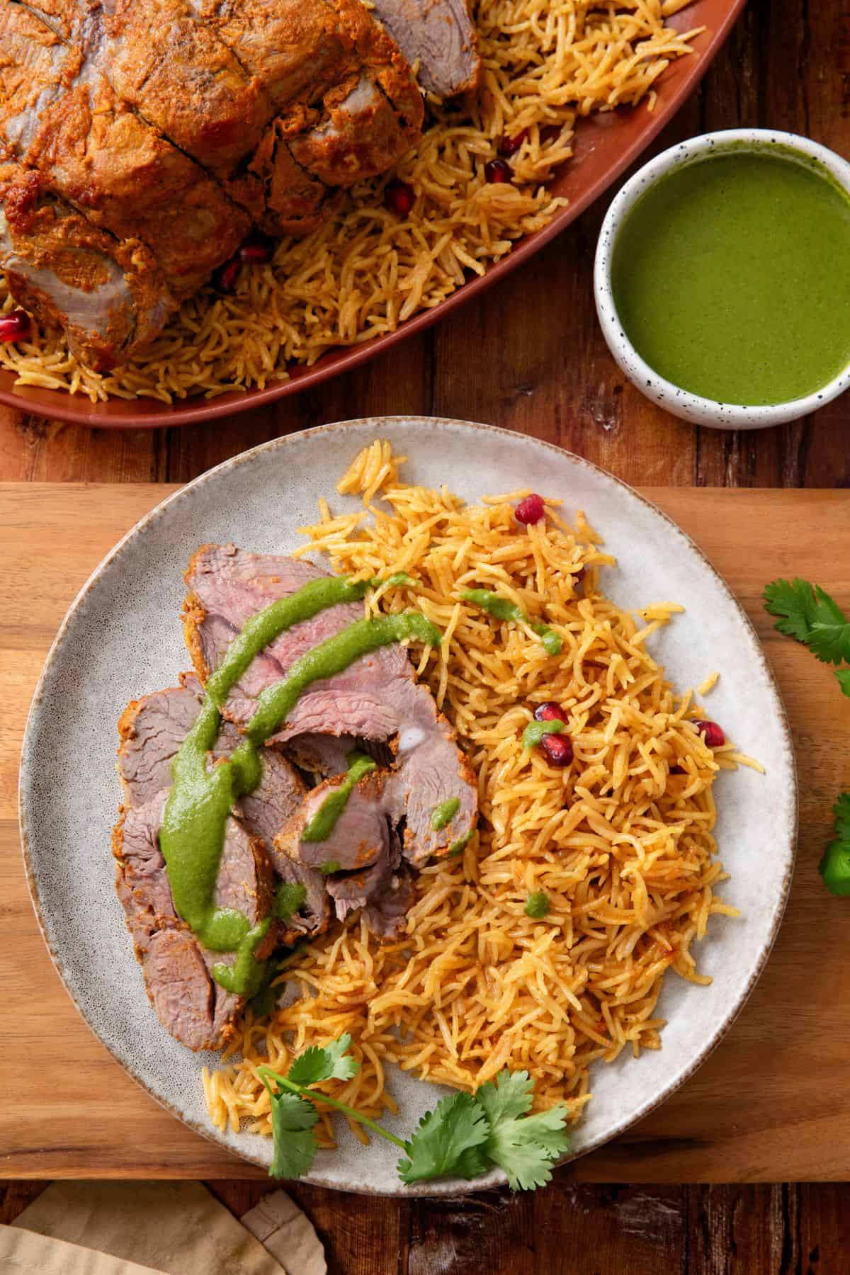 Sliced lamb roast is served in a white plate with rice and green chutney.