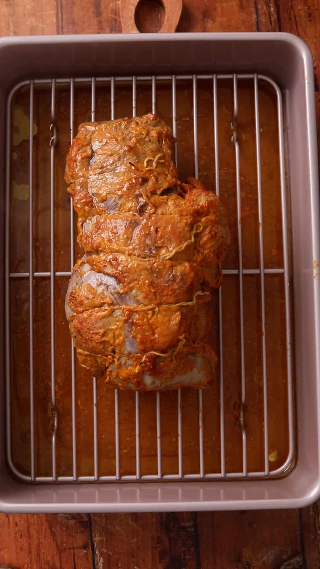 A roasted lamb leg sits on the metal rack inside a roasting pan with juices beneath the rack.